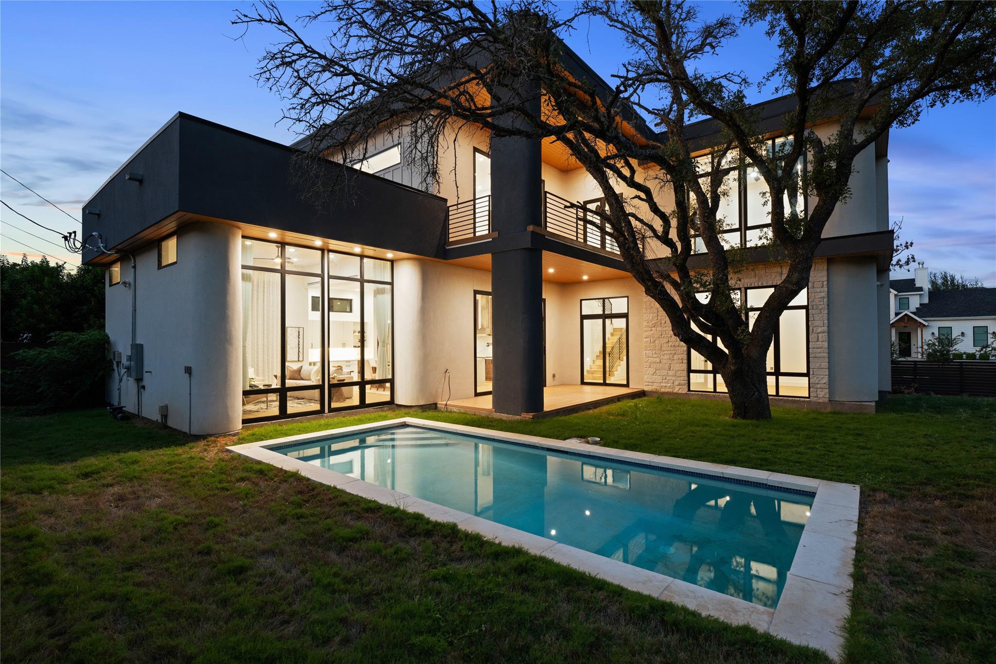 Modern two-story home and swimming pool at dusk, framed by a large tree.