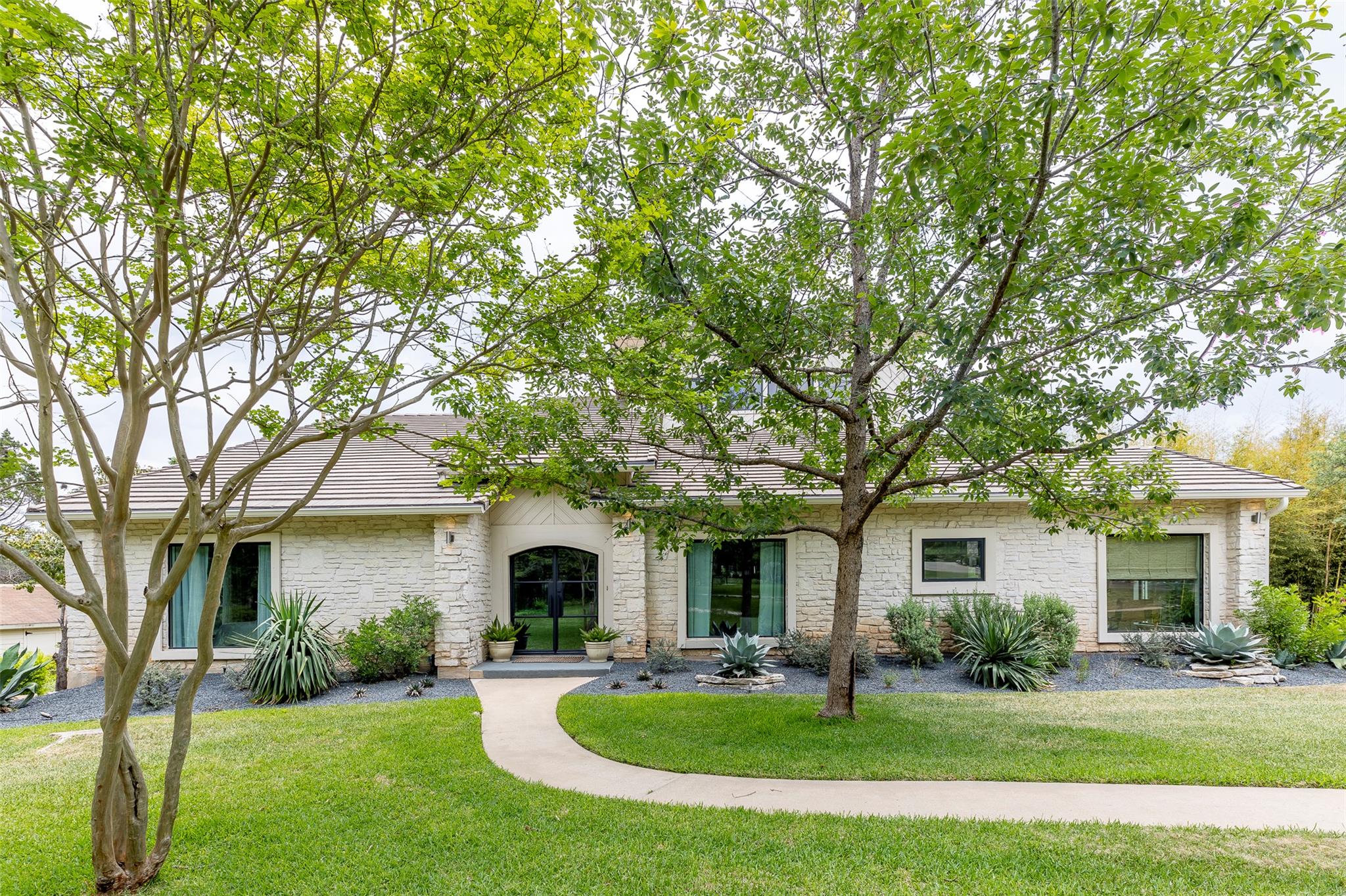 Single-story stone house framed by leafy trees, with a curved walkway and landscaped front yard.