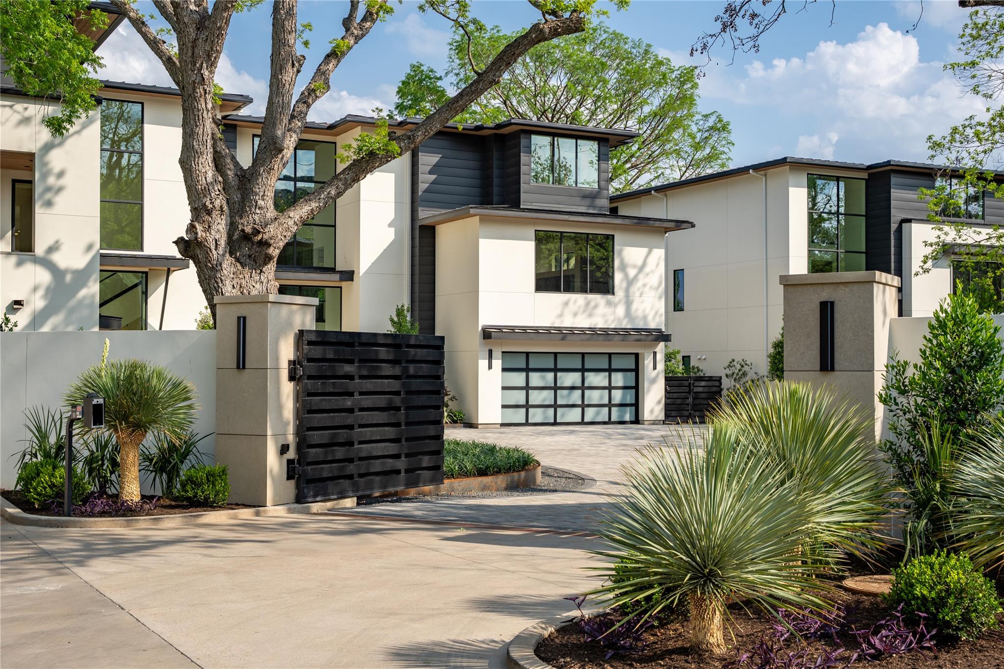 Contemporary two-story home with a black slatted gate, glass-paneled garage door, and mature trees.