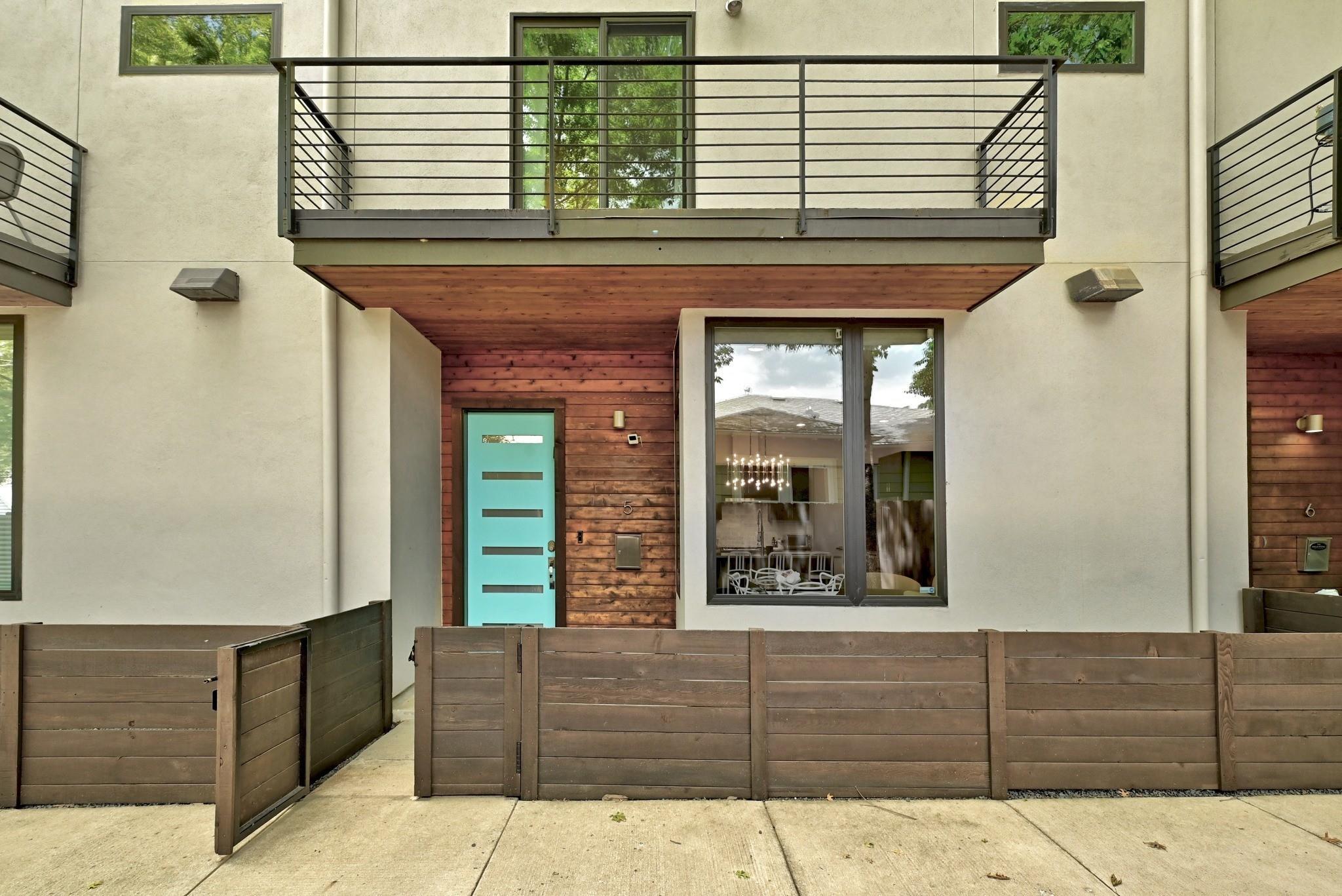Modern residence with a turquoise front door, wood accents, and a fenced patio beneath a balcony.