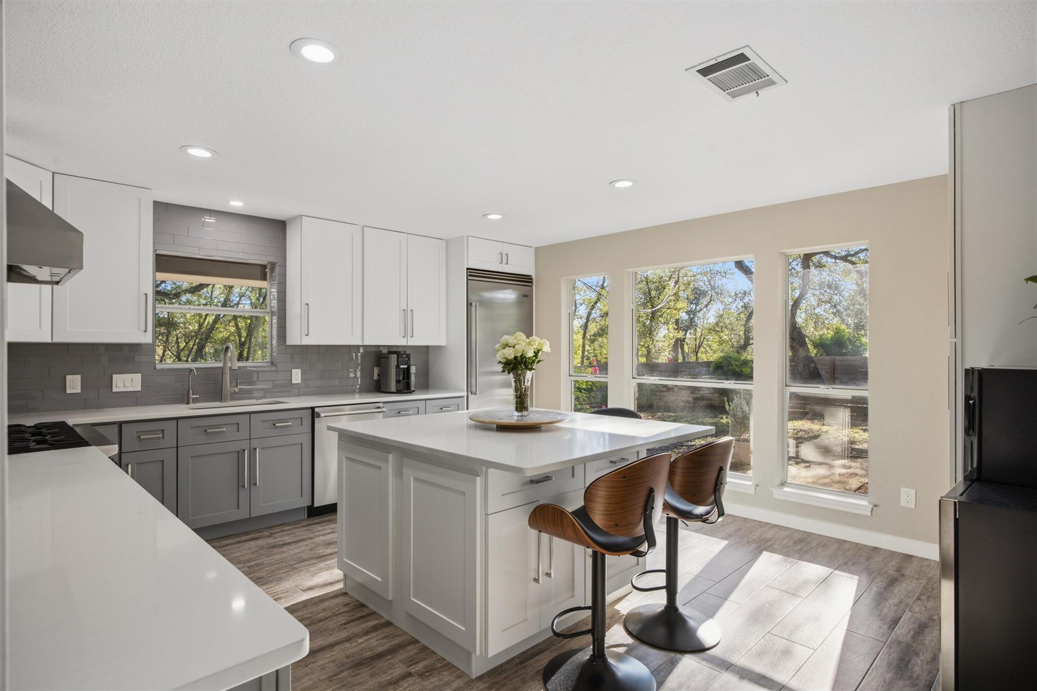 Bright modern kitchen with a large island, wood bar stools, and windows overlooking trees.