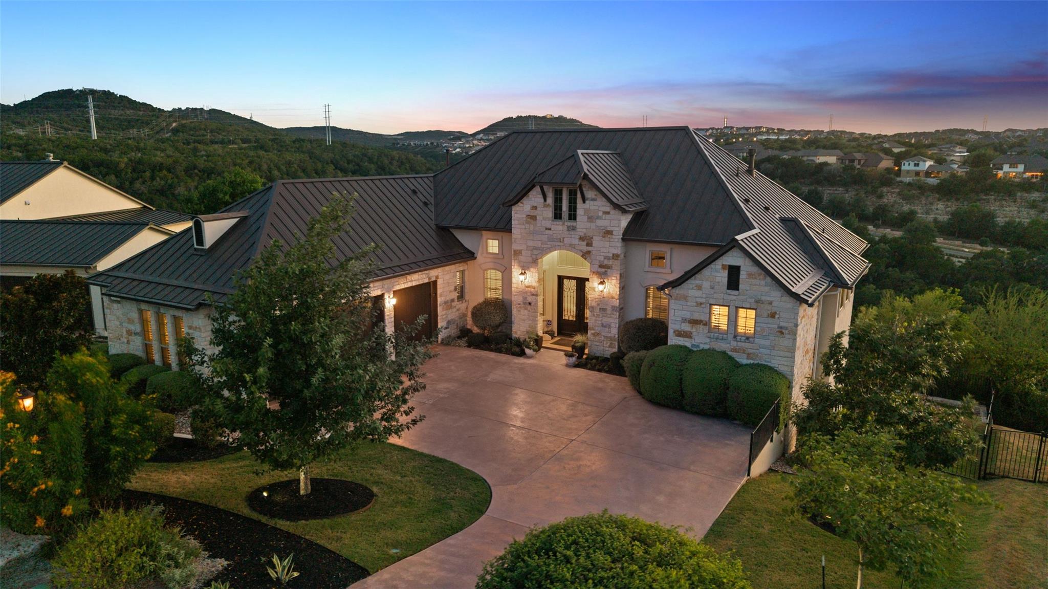 Stone house with dark metal roofs and landscaped driveway at dusk, backed by wooded hills.