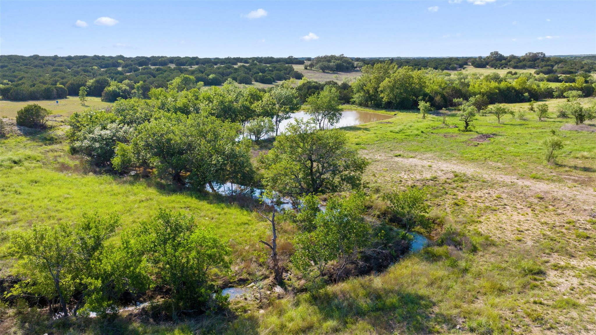 Aerial view of grassy rural land with scattered trees, a pond, and a winding creek.