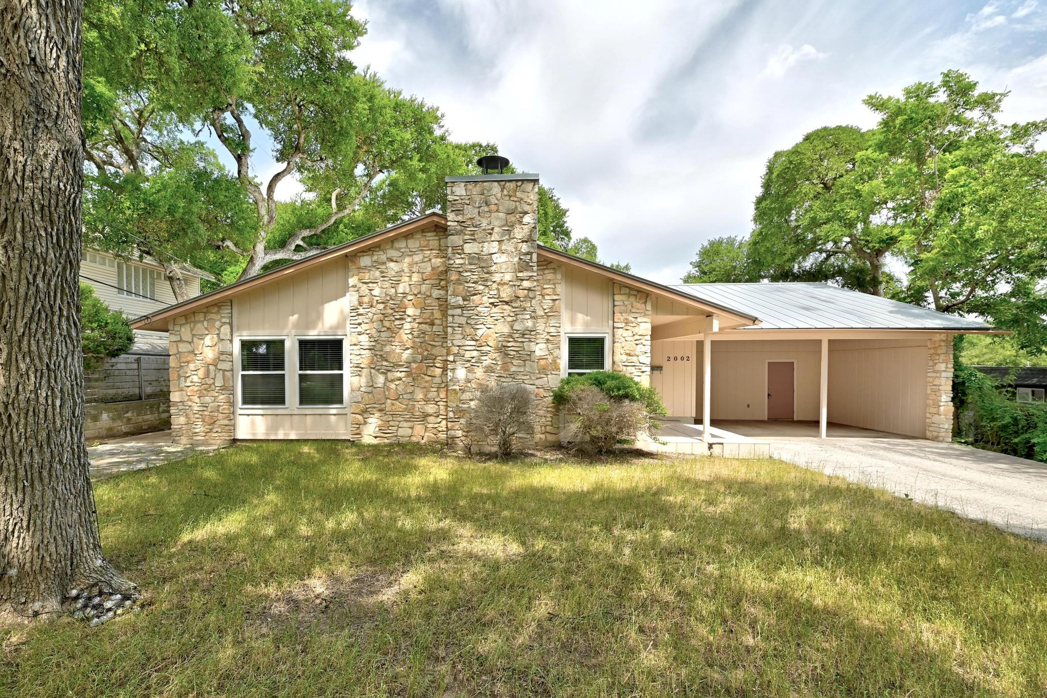 Single-story stone house with a tall chimney, covered carport, and mature trees.