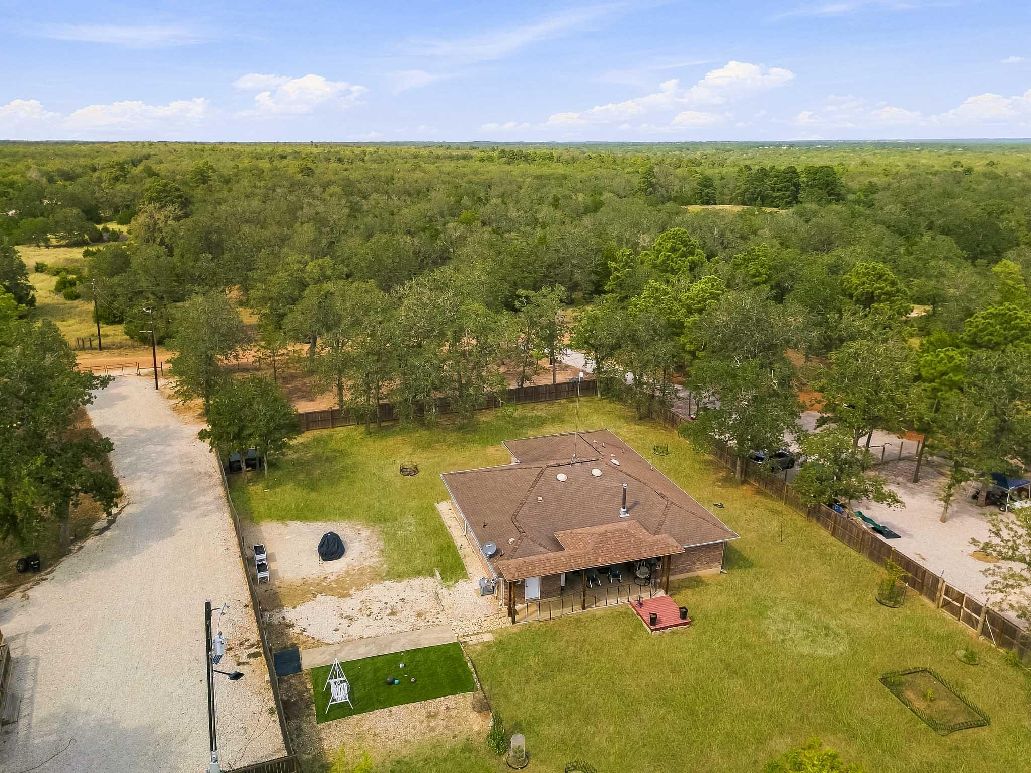 Aerial view of a single-story home with a fenced lawn, gravel driveway, and surrounding woodland.