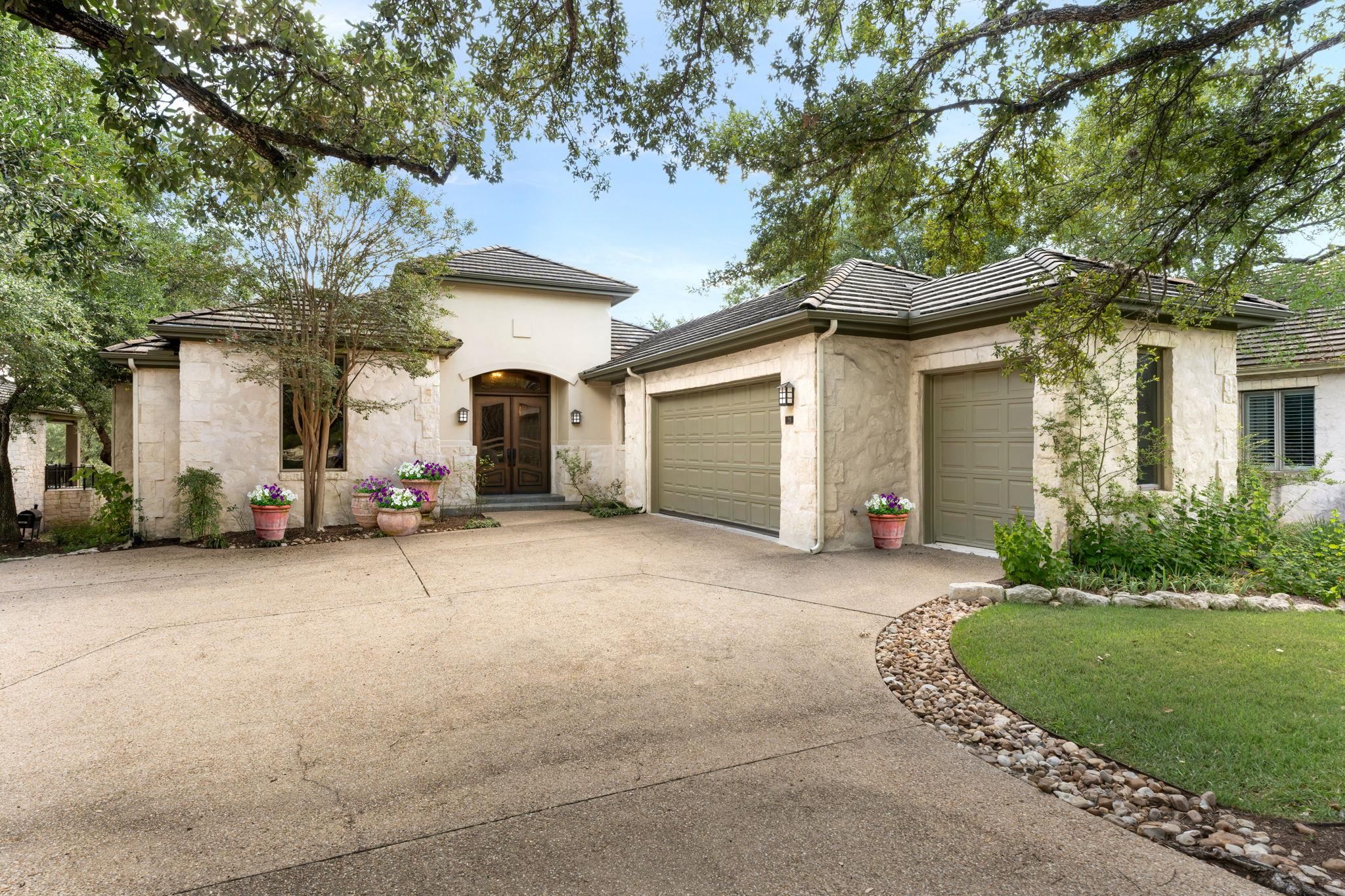 Stone home with two garage doors, a broad driveway, potted flowers, and mature trees.