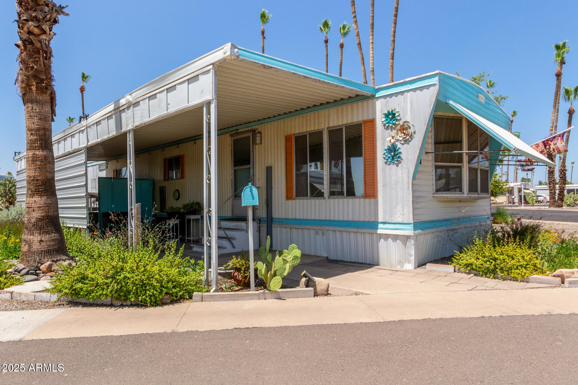 White manufactured home with turquoise trim, a covered carport, orange shutters, and palm trees.