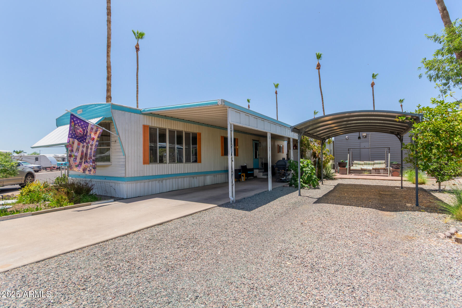 Manufactured home with turquoise trim, covered porch, and a metal carport over gravel parking.