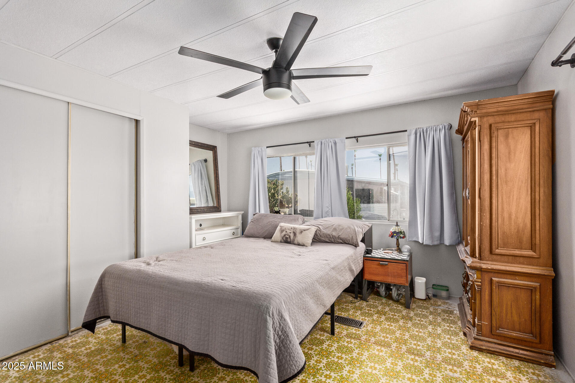 Furnished bedroom with a large bed, patterned carpet, ceiling fan, and tall wooden armoire.