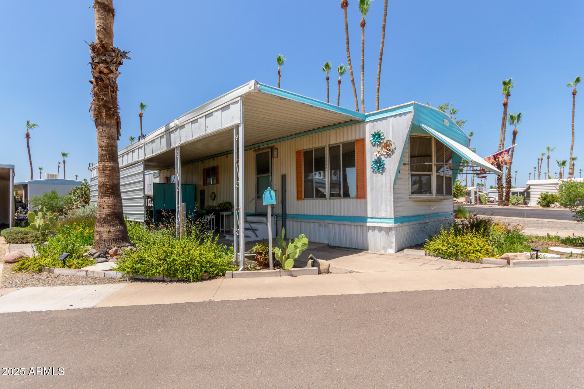 White manufactured home with turquoise trim and a covered carport, surrounded by palms and cacti.