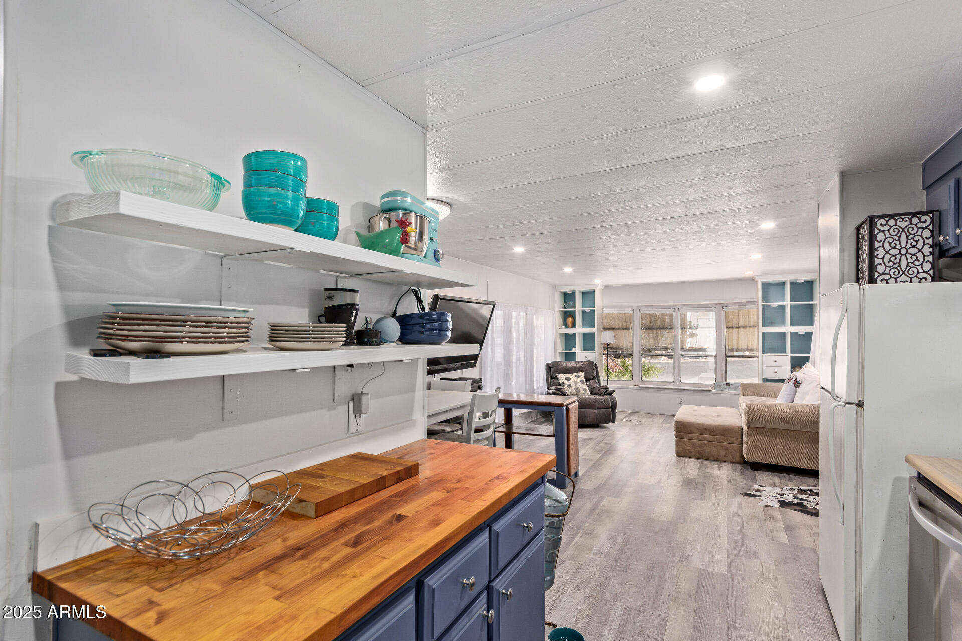 Open-plan kitchen with blue cabinets, butcher-block counters, and open shelves, looking toward a bright living area.