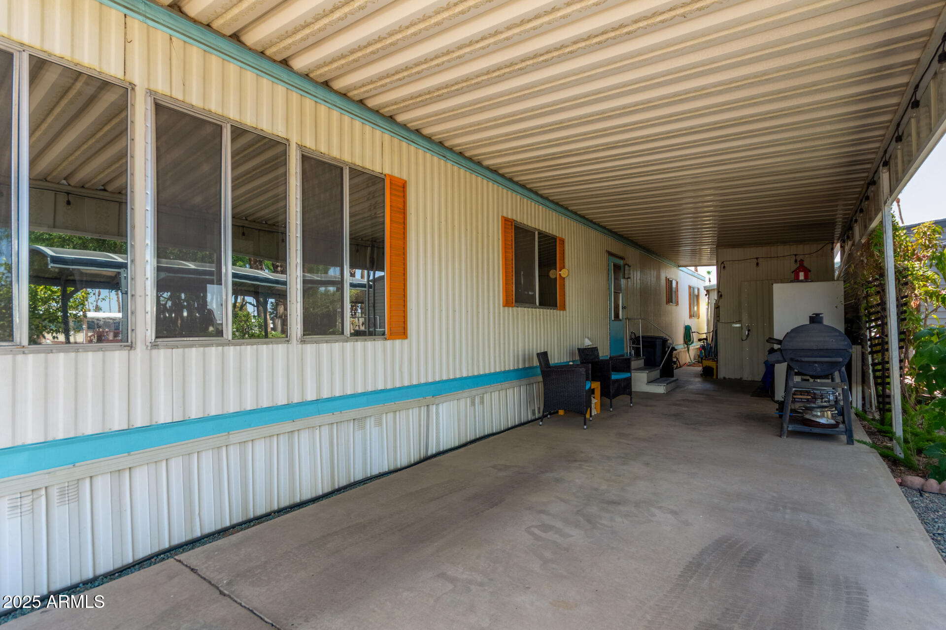 Covered patio beside a manufactured home, with turquoise trim, outdoor chairs, and a smoker grill.