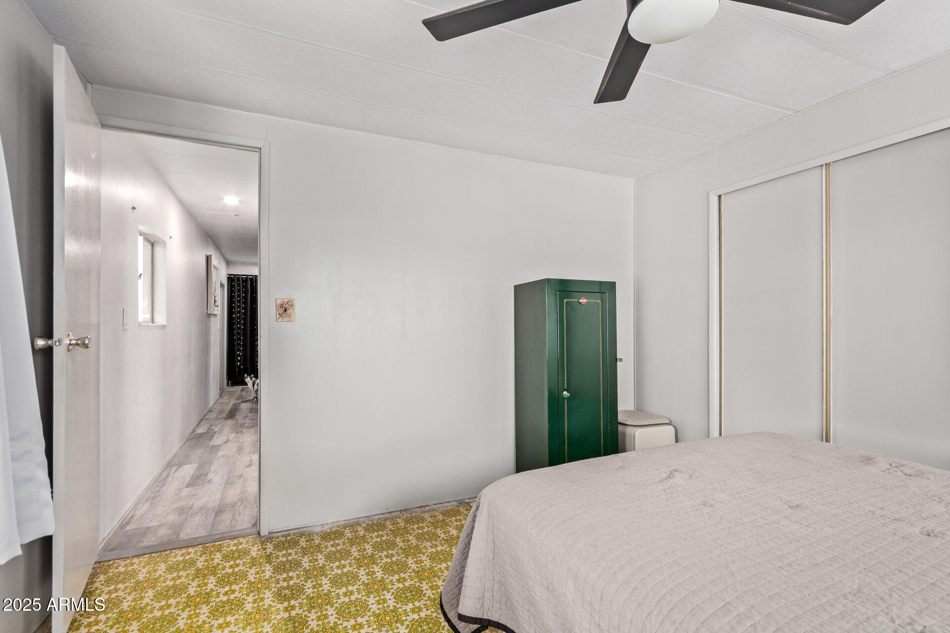 Bedroom with a white bed, green safe, ceiling fan, and patterned flooring beside a hallway.
