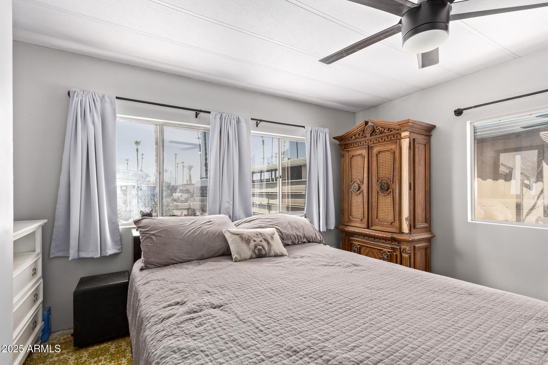 Bedroom with a gray quilted bed, wide curtained windows, and an ornate wooden armoire.