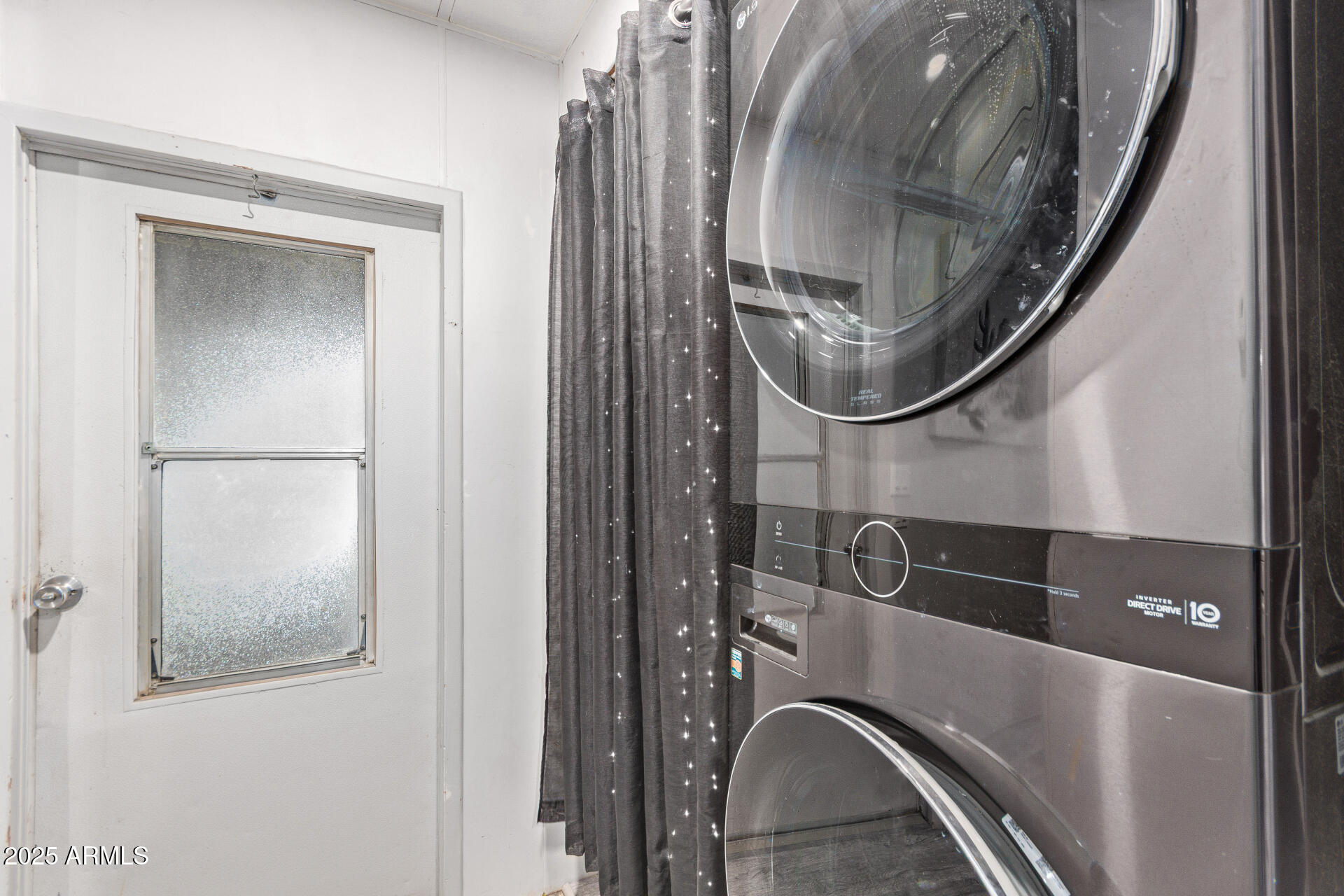 Stacked front-loading washer and dryer beside a frosted-glass door and dark curtain.