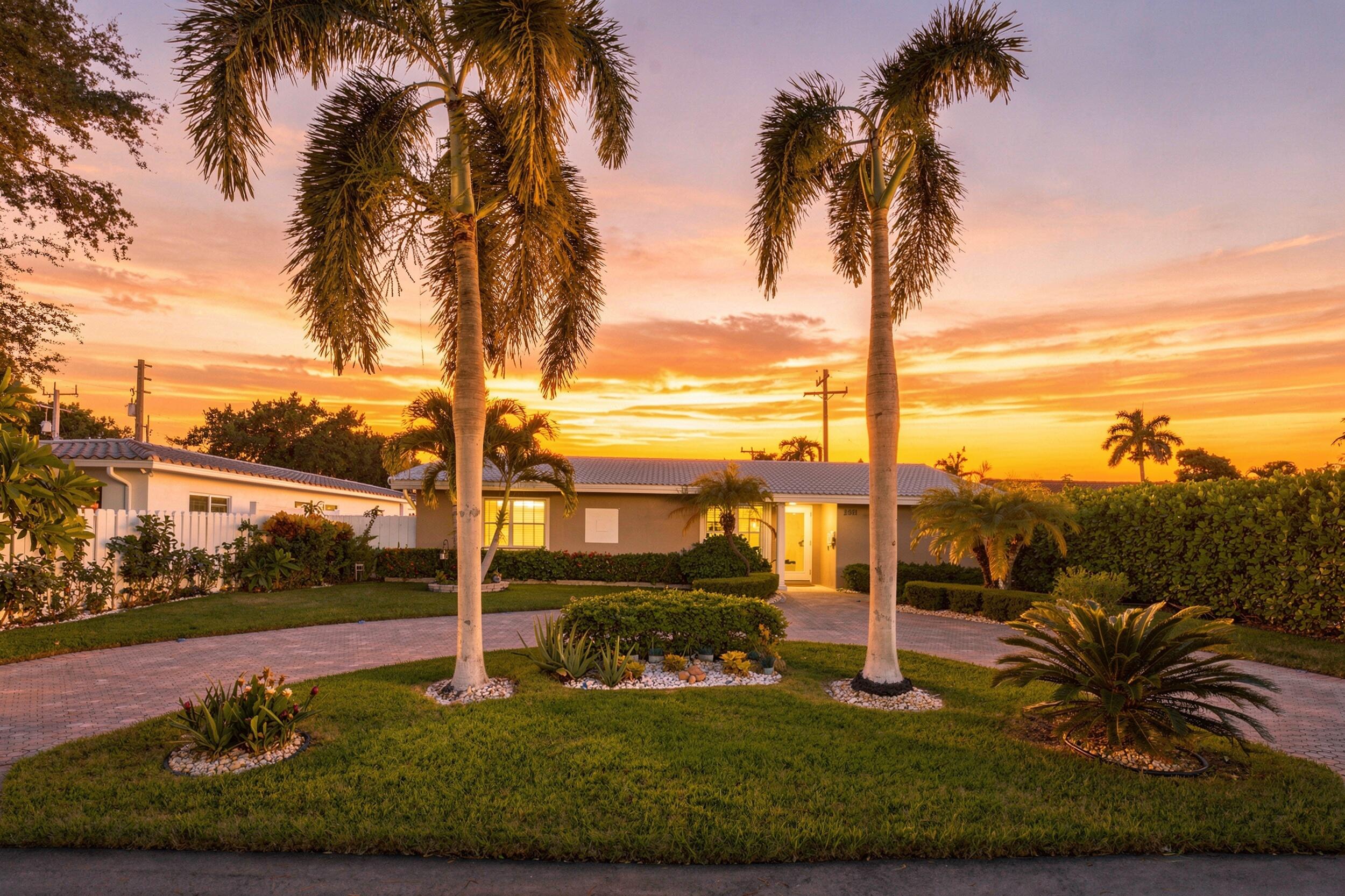 Single-story home framed by tall palms, with a landscaped lawn and paver driveway at sunset.