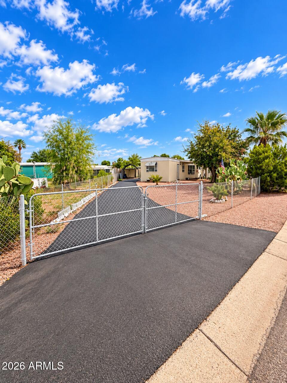 Fenced property entrance with a paved driveway leading to a beige home amid desert landscaping.