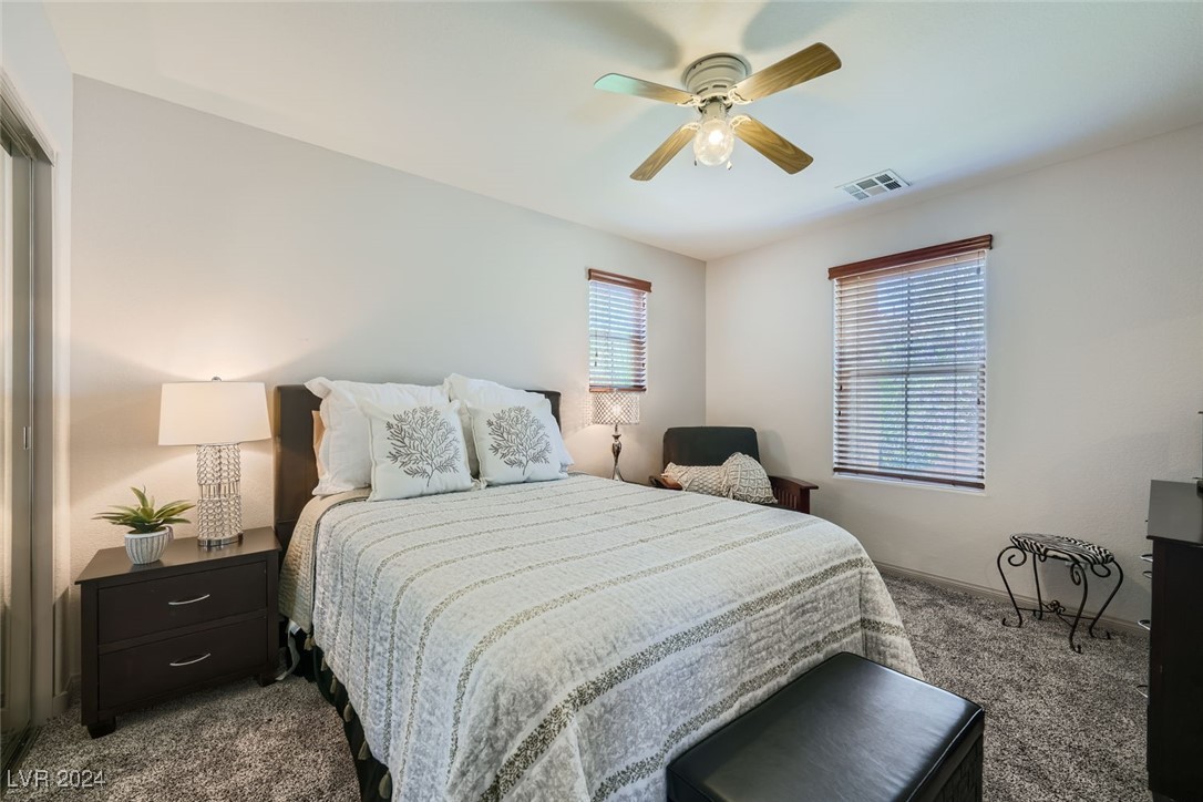 Furnished bedroom with a pale quilted bed, two blinded windows, and a ceiling fan.