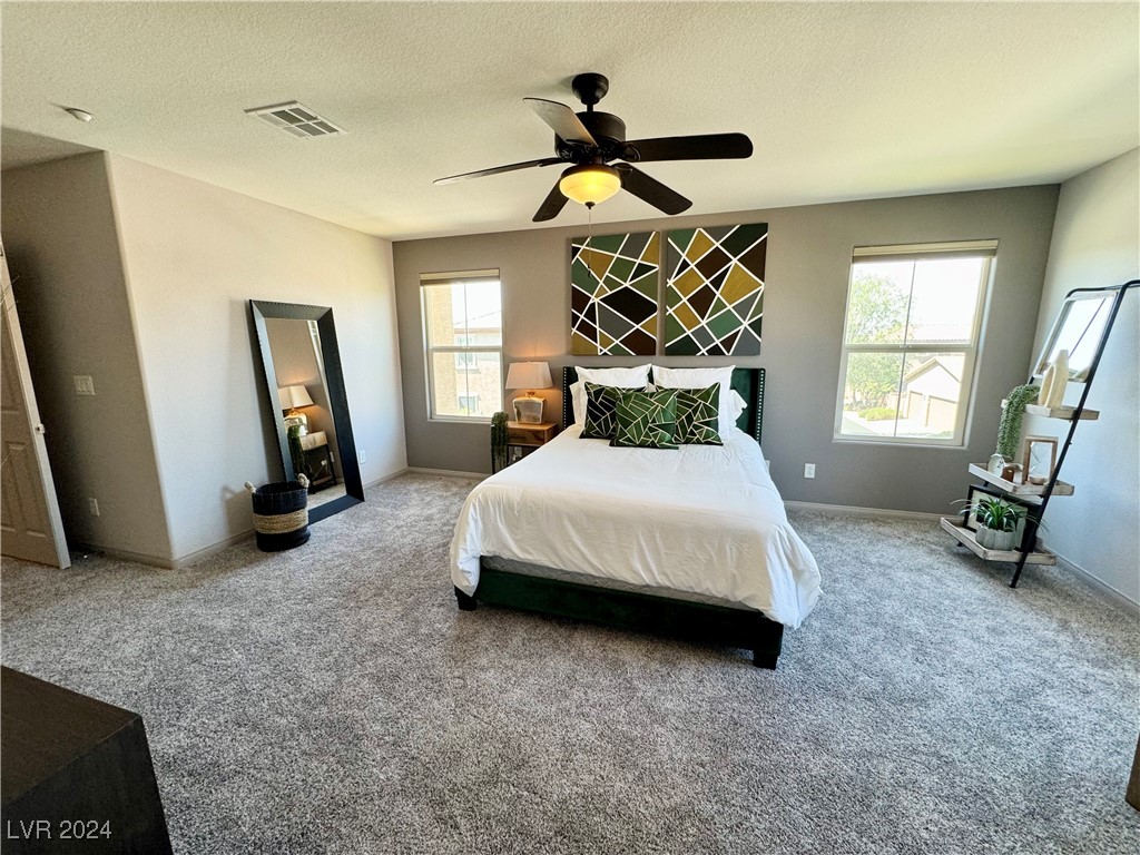 Staged bedroom with a king bed, geometric green artwork, gray carpet, and ceiling fan.