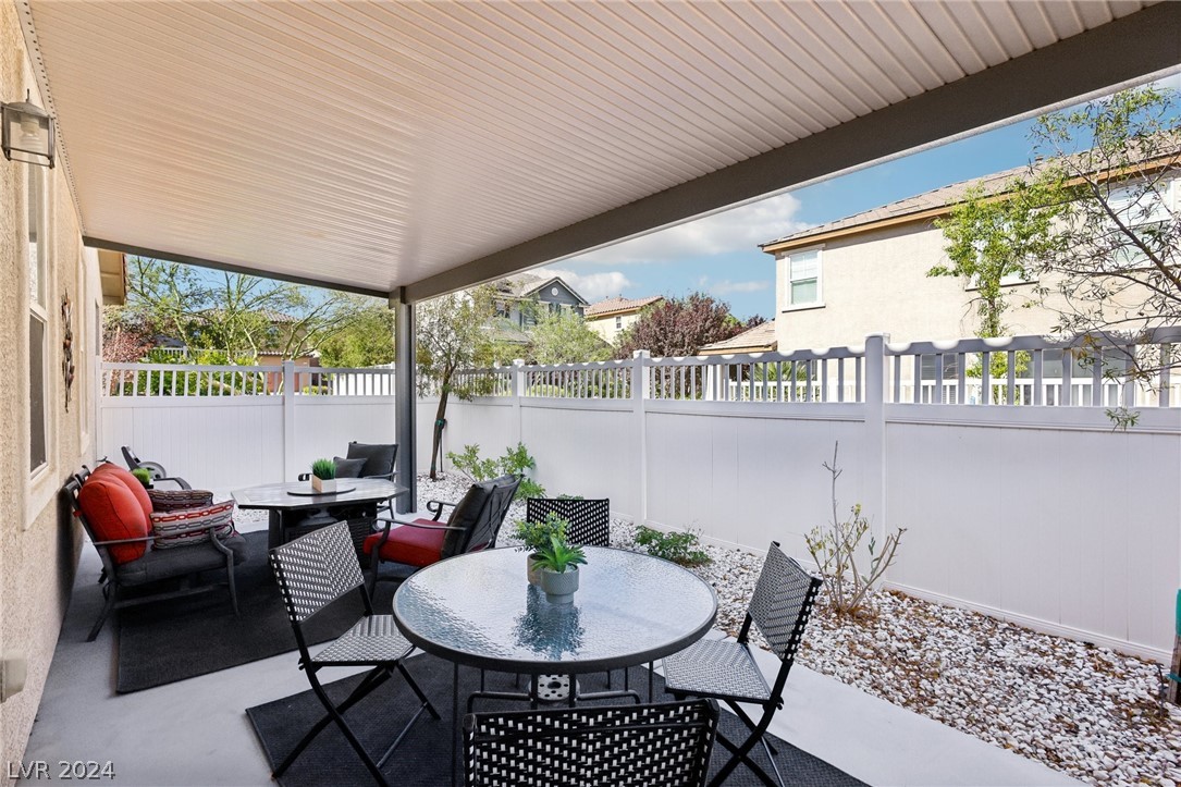 Covered backyard patio with glass-top dining table, red-cushioned lounge seating, and white privacy fence.