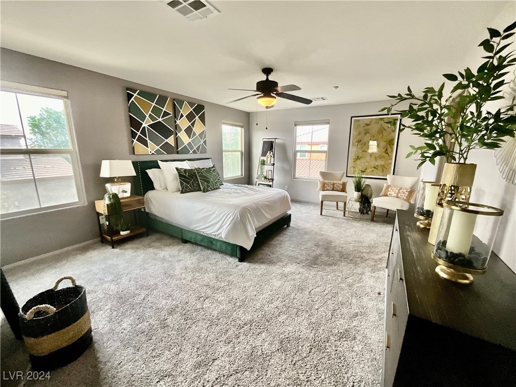 Staged bedroom with a white bed, geometric wall art, gray carpet, and a sitting area by the windows.