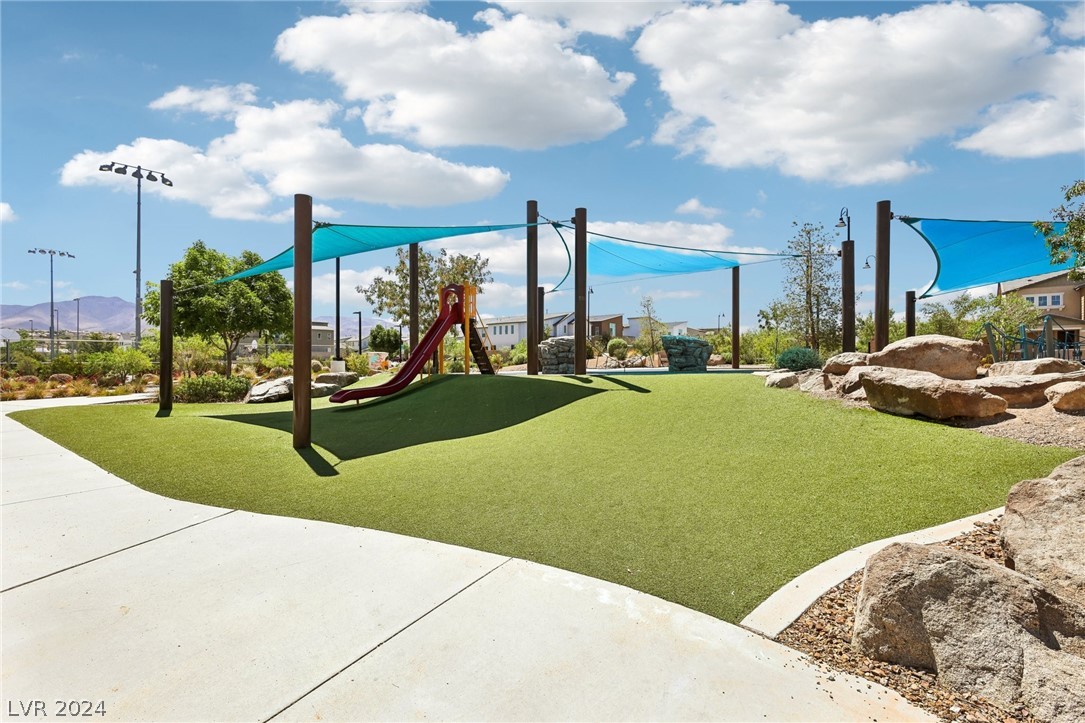 Community playground with a red slide, artificial turf, turquoise shade sails, and distant mountains.