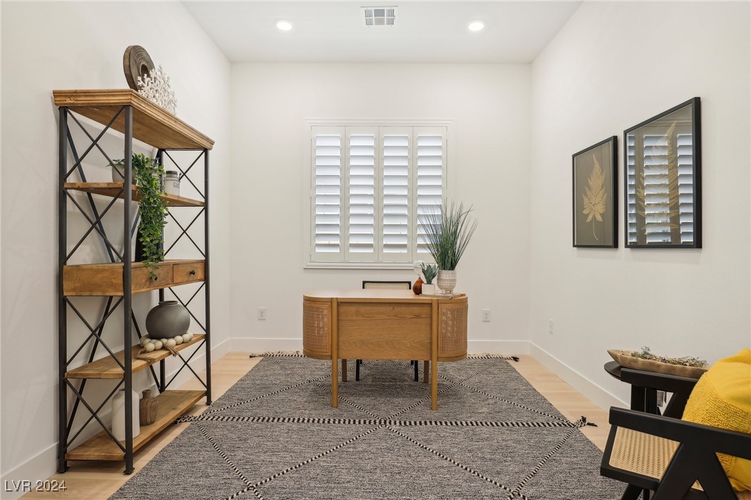 Staged home office with a wooden desk, shuttered window, patterned rug, and open display shelving.