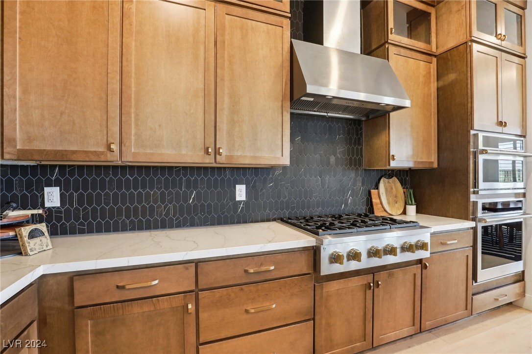 Kitchen with warm wood cabinets, dark geometric tile backsplash, white counters, and stainless-steel appliances.