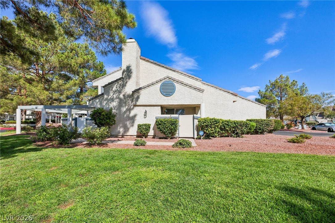 Light stucco building with a gabled roof and round vent, surrounded by shrubs, lawn, and pine trees.