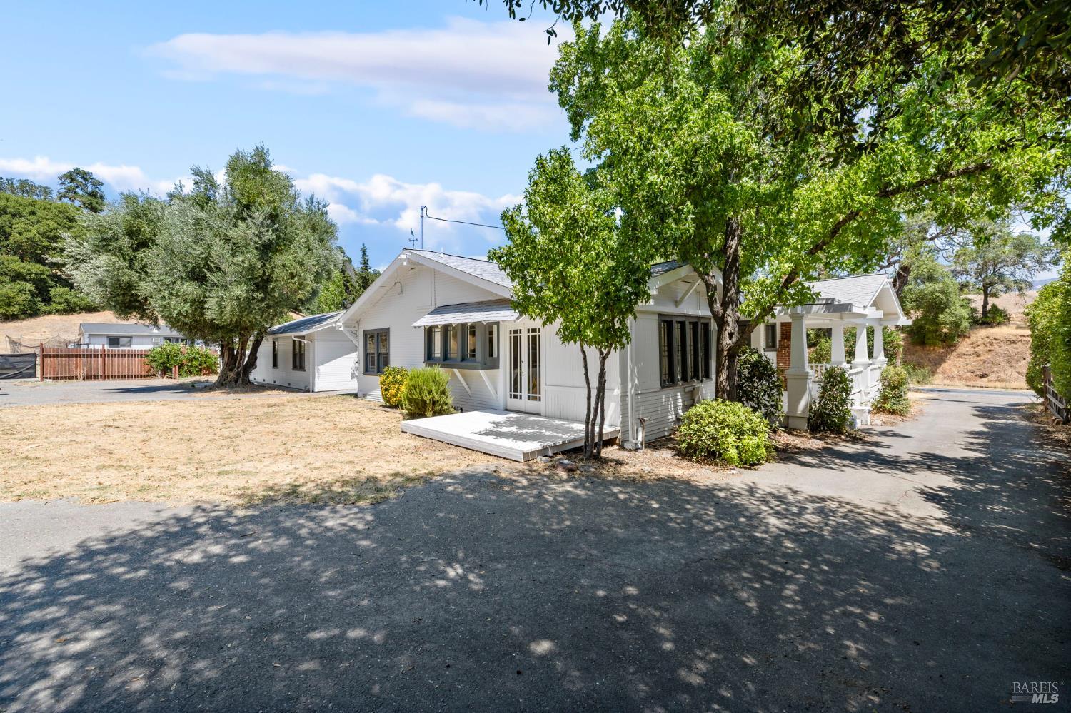 White single-story house with a covered porch, front deck, and mature trees.