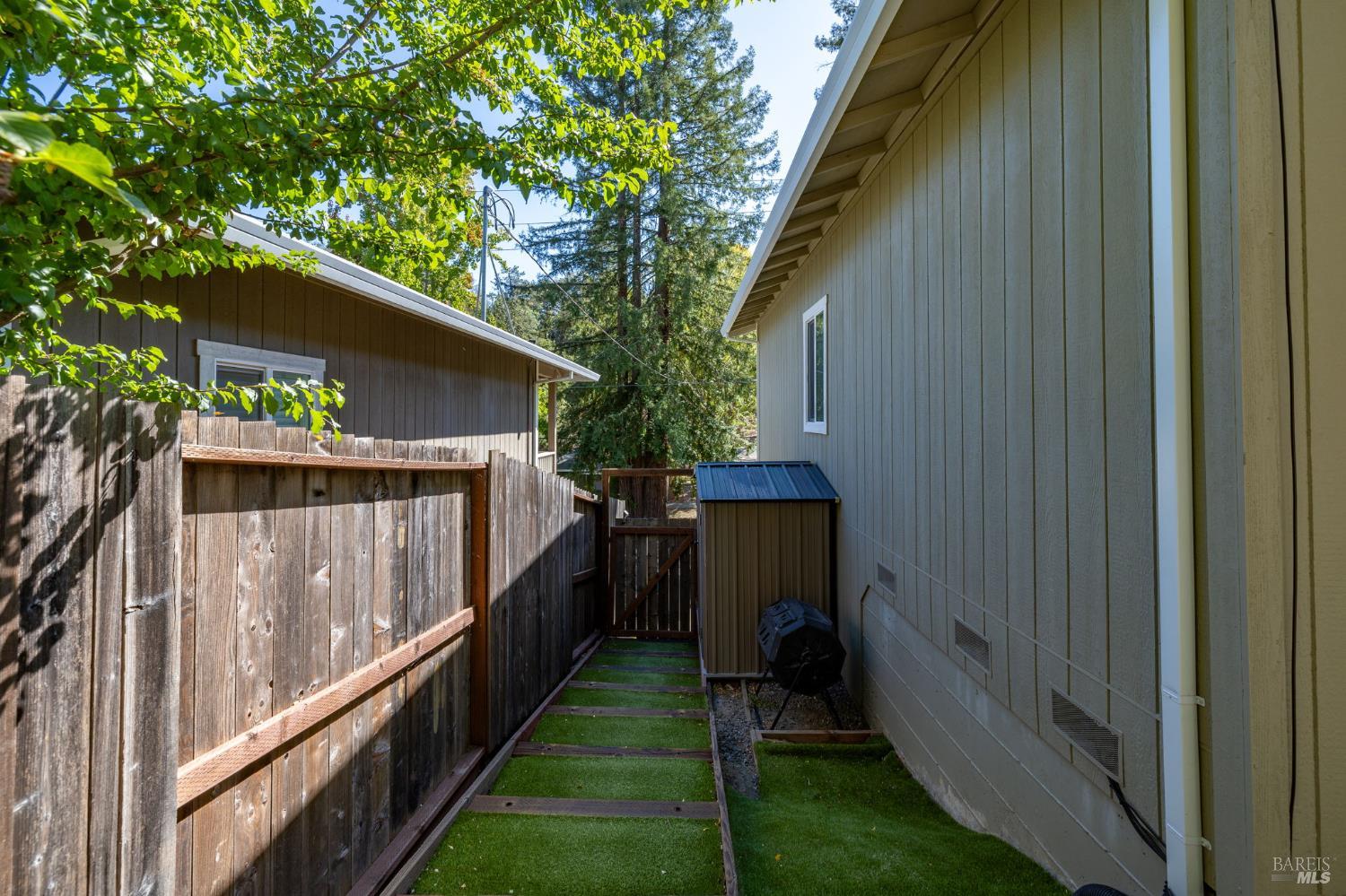 Side yard with artificial turf between a gray-sided house and wooden fence, leading to a shed.