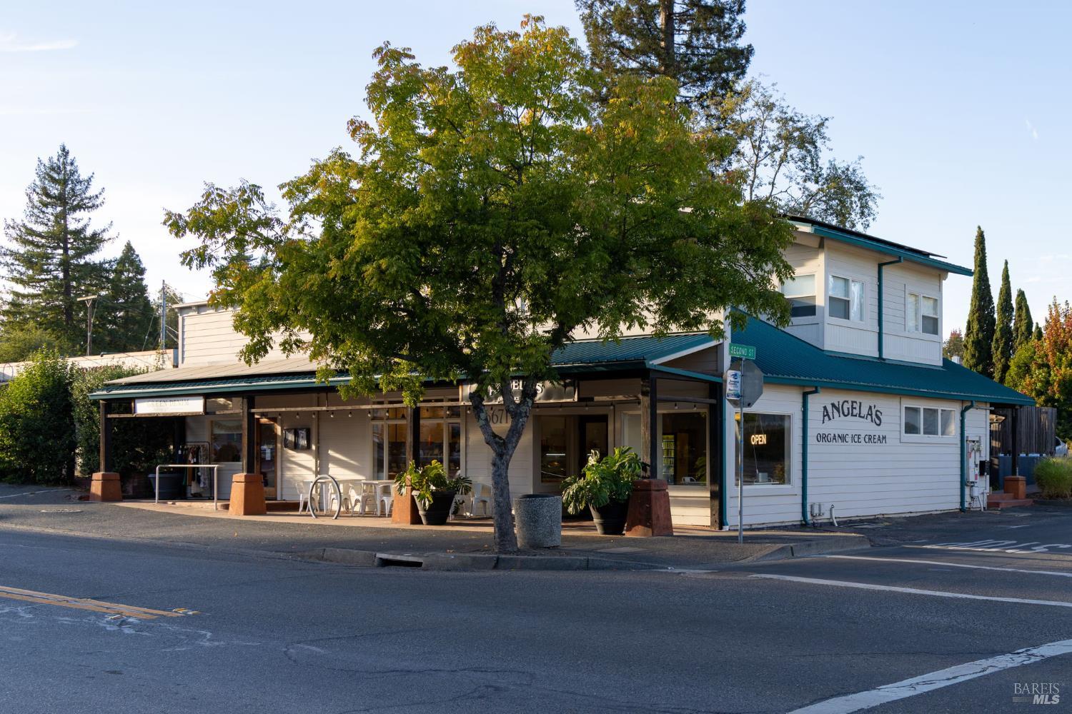Two-story white storefront with teal awnings, partly shaded by a large tree.