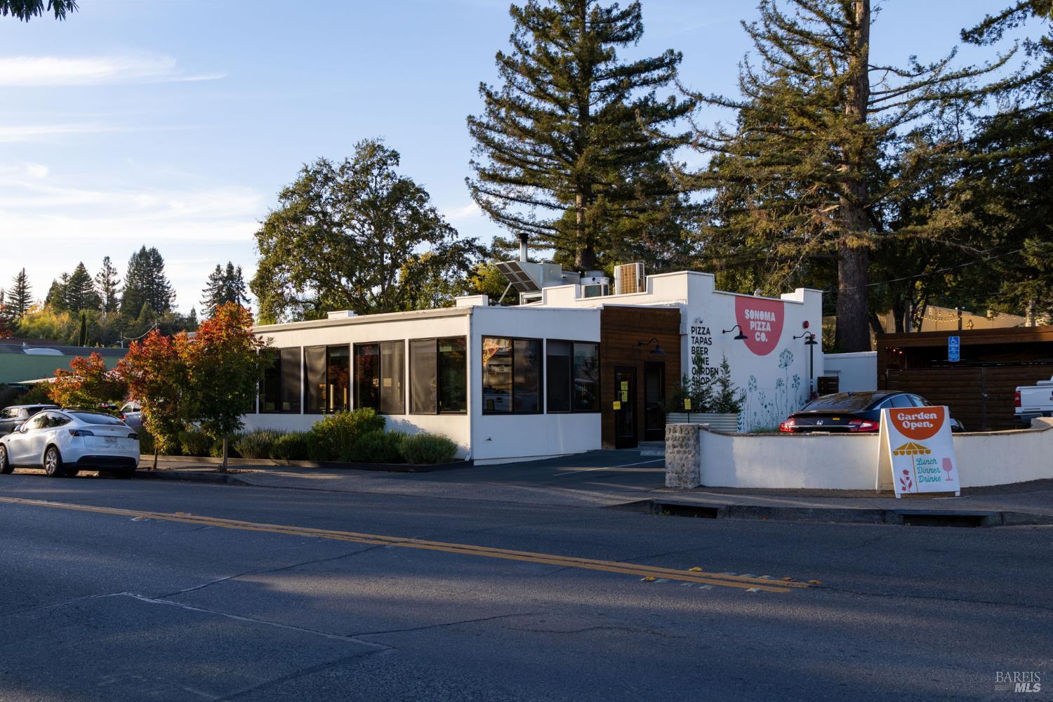 White Sonoma Pizza Co. restaurant with broad windows, a dark wood entrance, and a garden-open sidewalk sign.