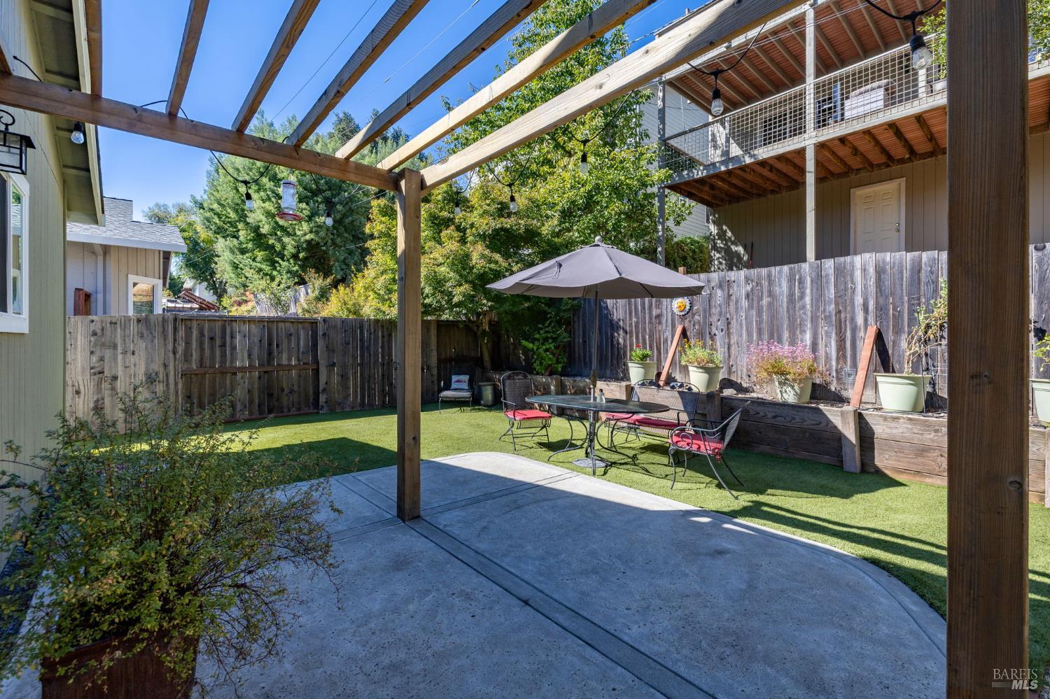 Backyard patio with a timber pergola, outdoor dining set, umbrella, artificial turf, and raised planters.