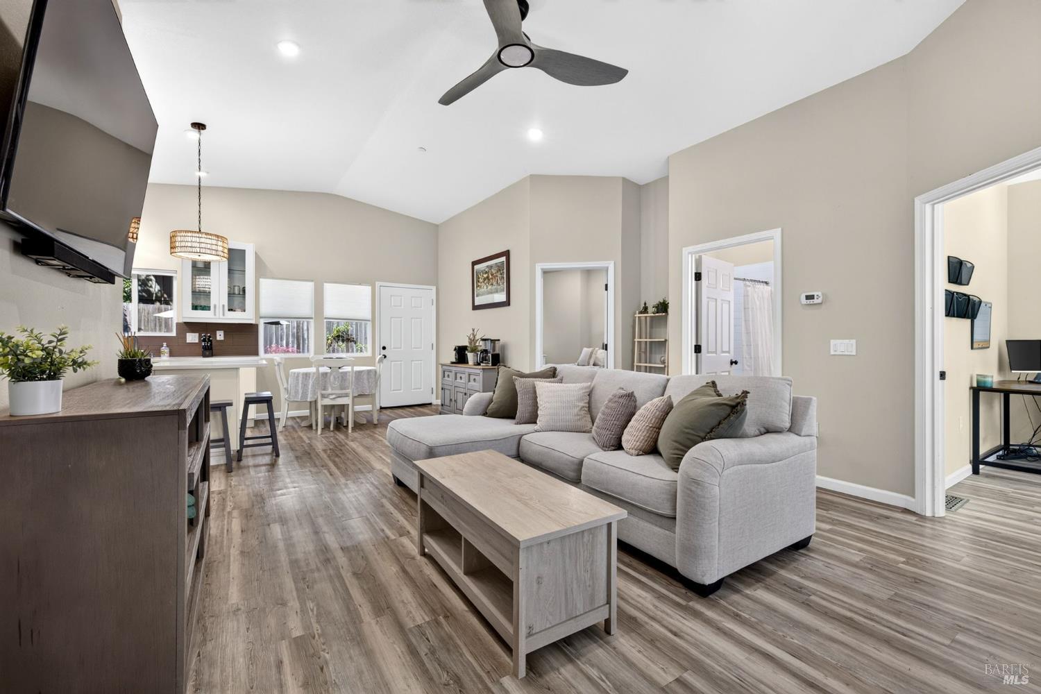 Open-plan living room with vaulted ceiling, gray sectional, wood floors, and adjoining kitchen.