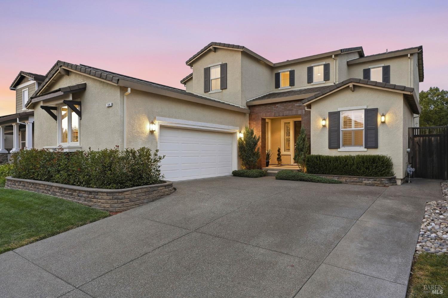Two-story stucco home with a brick entry, attached garage, landscaped beds, and a broad driveway at dusk.