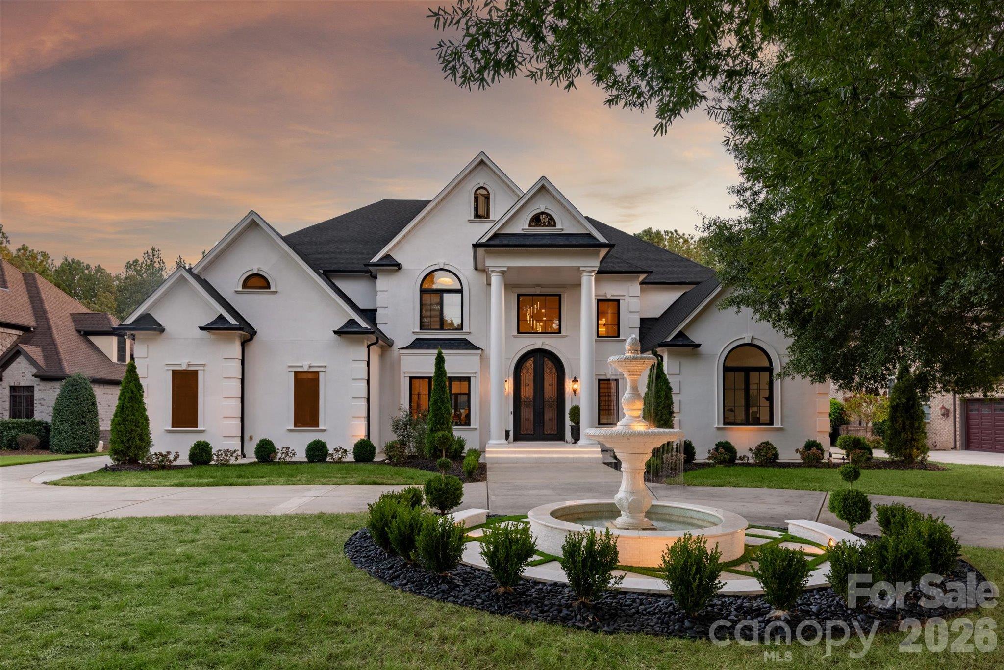 White two-story home with a columned entrance and tiered fountain at dusk.