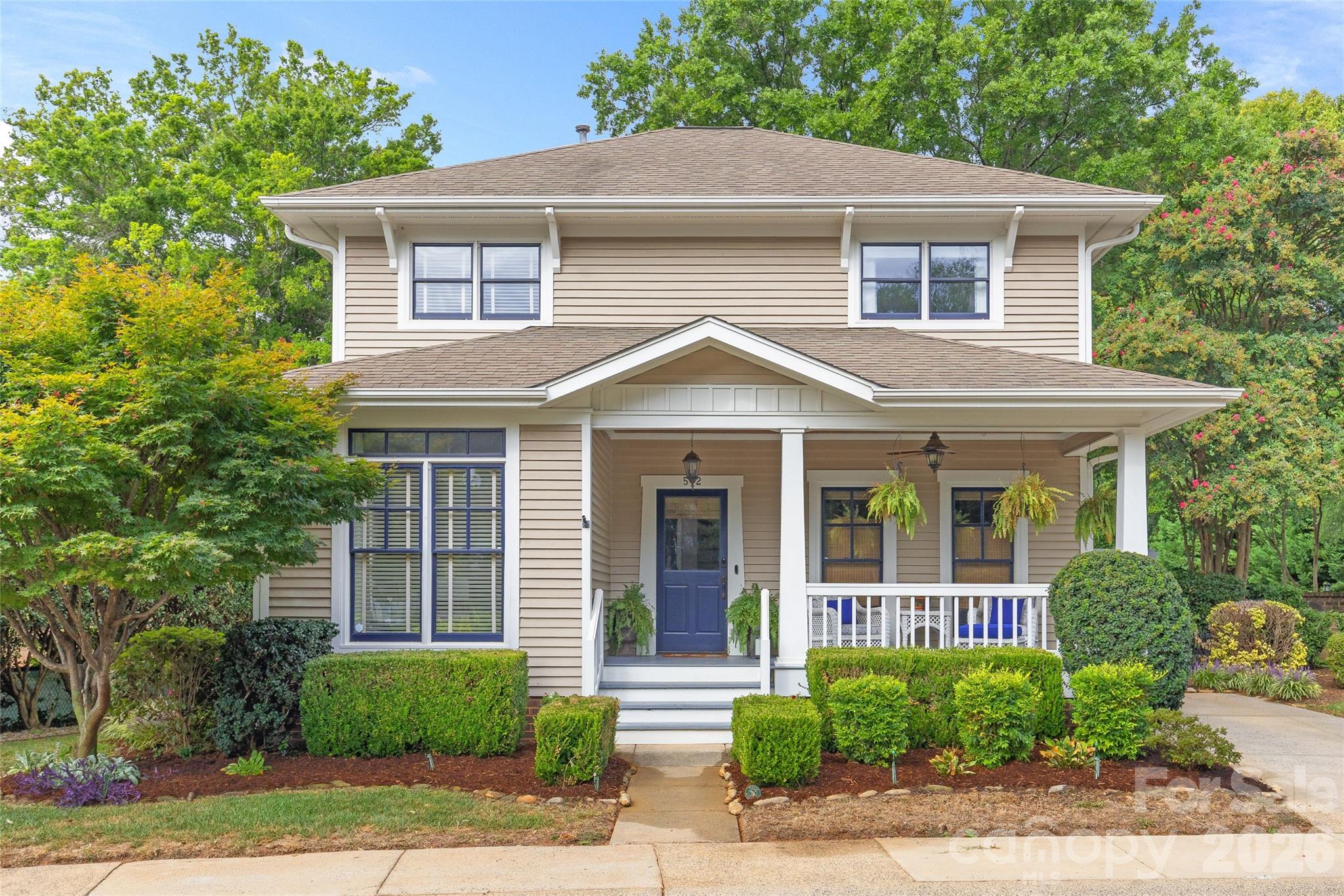 Two-story beige home with a blue front door, covered porch, hanging ferns, and neatly trimmed shrubs.