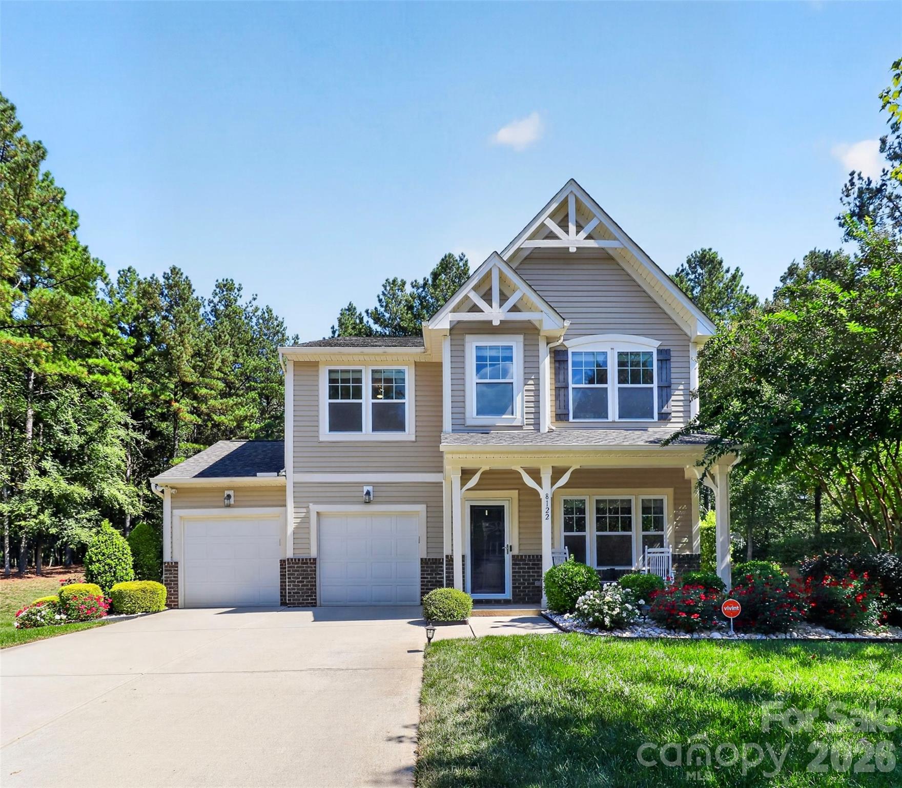 Two-story beige home with white gables, a covered porch, and a two-car garage, surrounded by trees and flower beds.