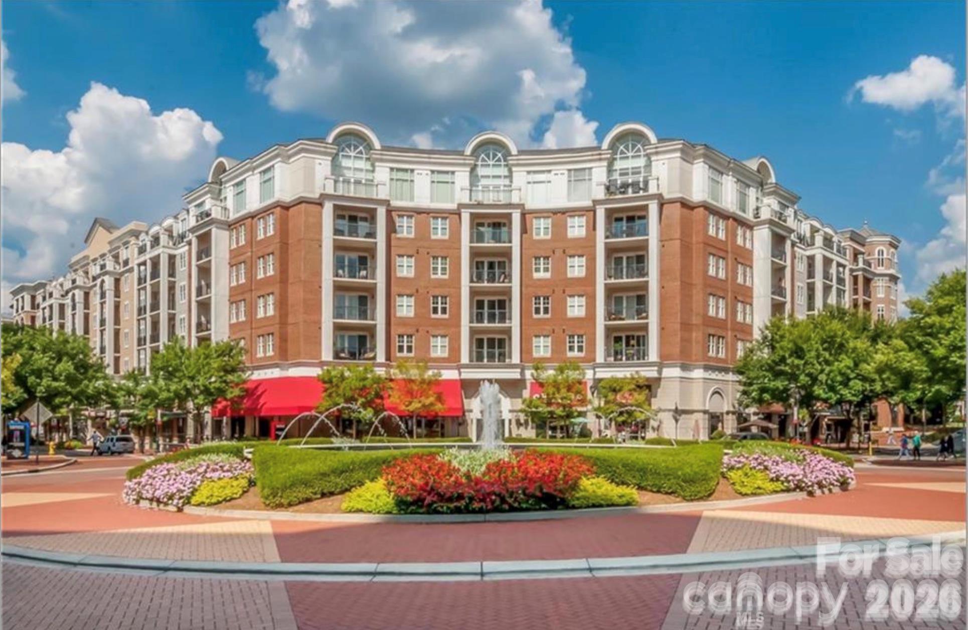 Brick condominium building with red awnings behind a landscaped plaza and fountain.