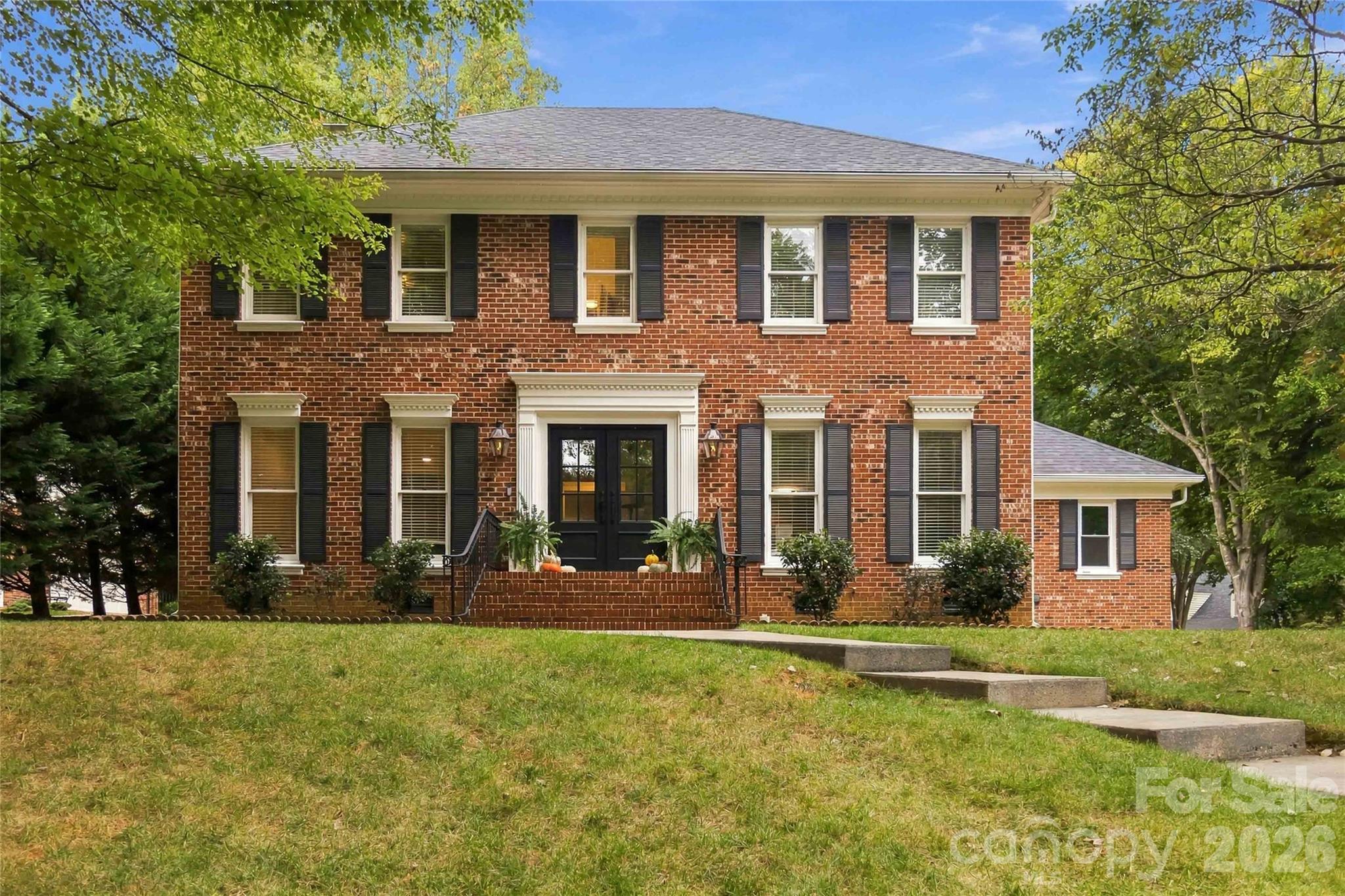 Two-story brick house with black shutters, white-trimmed windows, and steps across a sloping lawn.