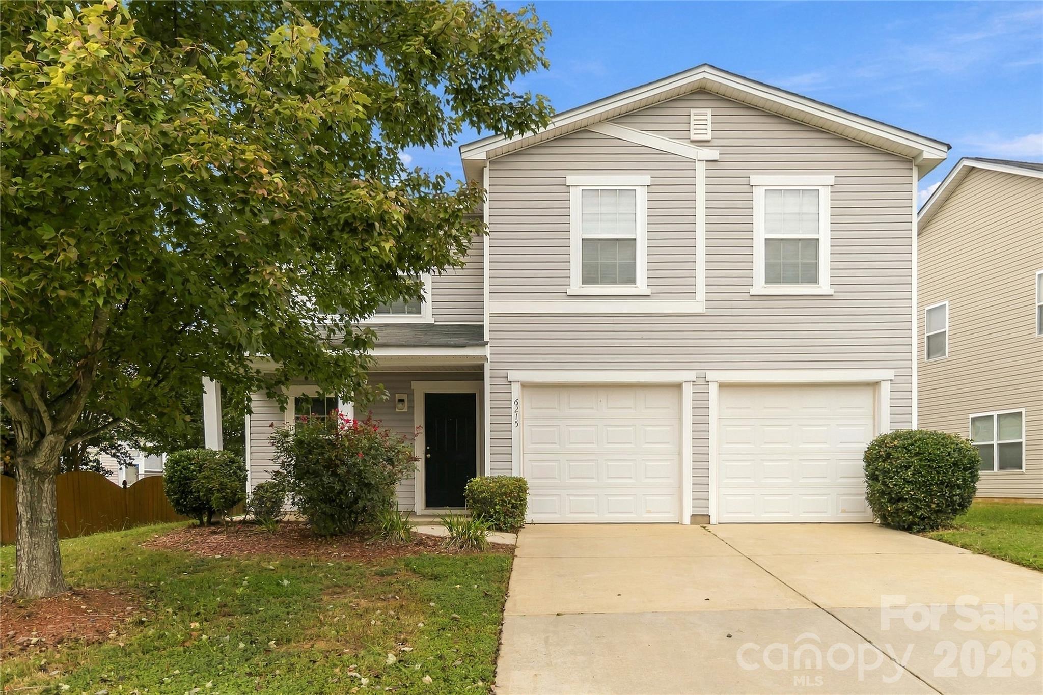 Two-story beige house with a double garage, broad driveway, and large leafy tree in front.