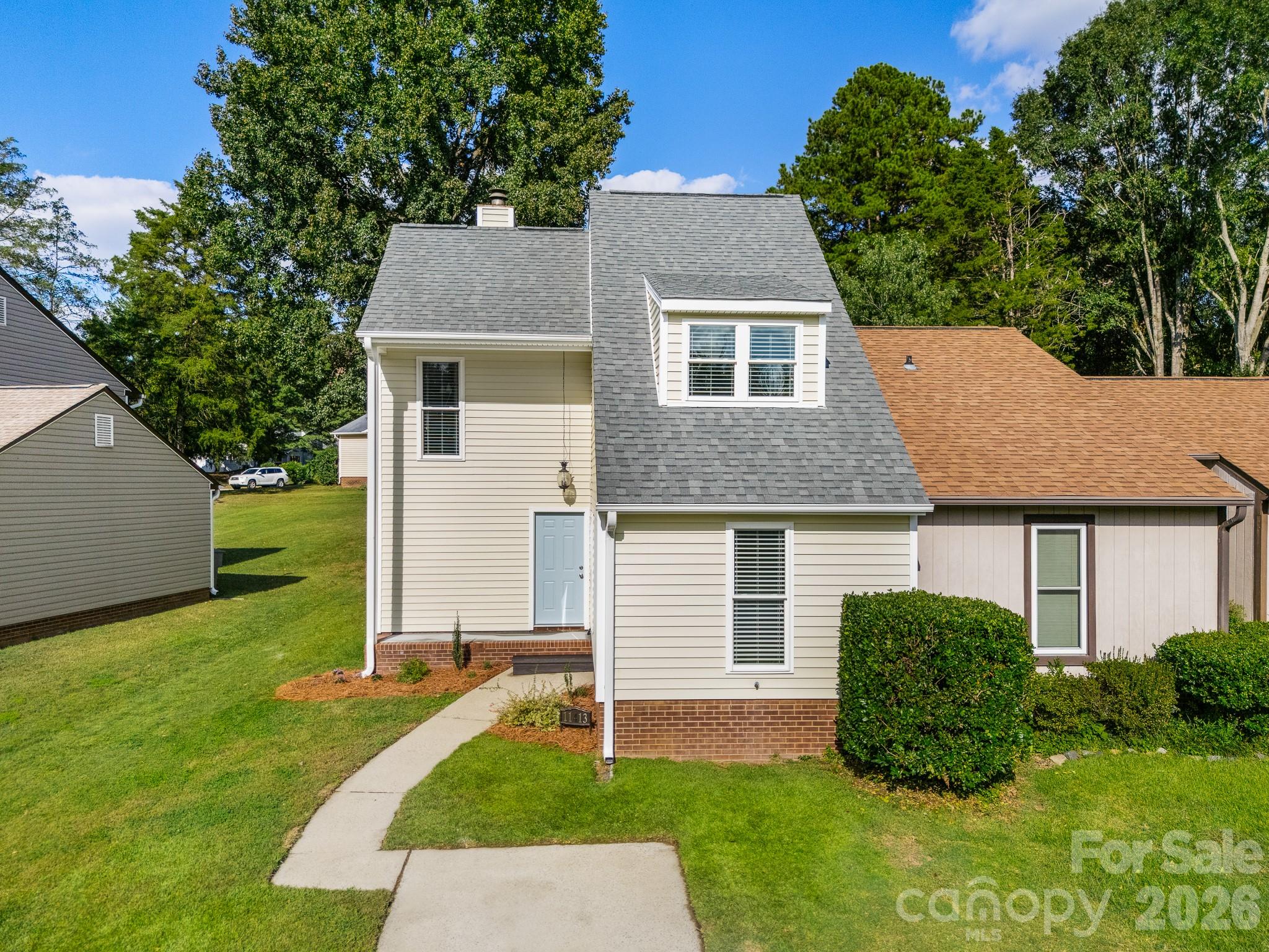 Beige two-story house with a gray roof, dormer, brick foundation, and curved front walkway.