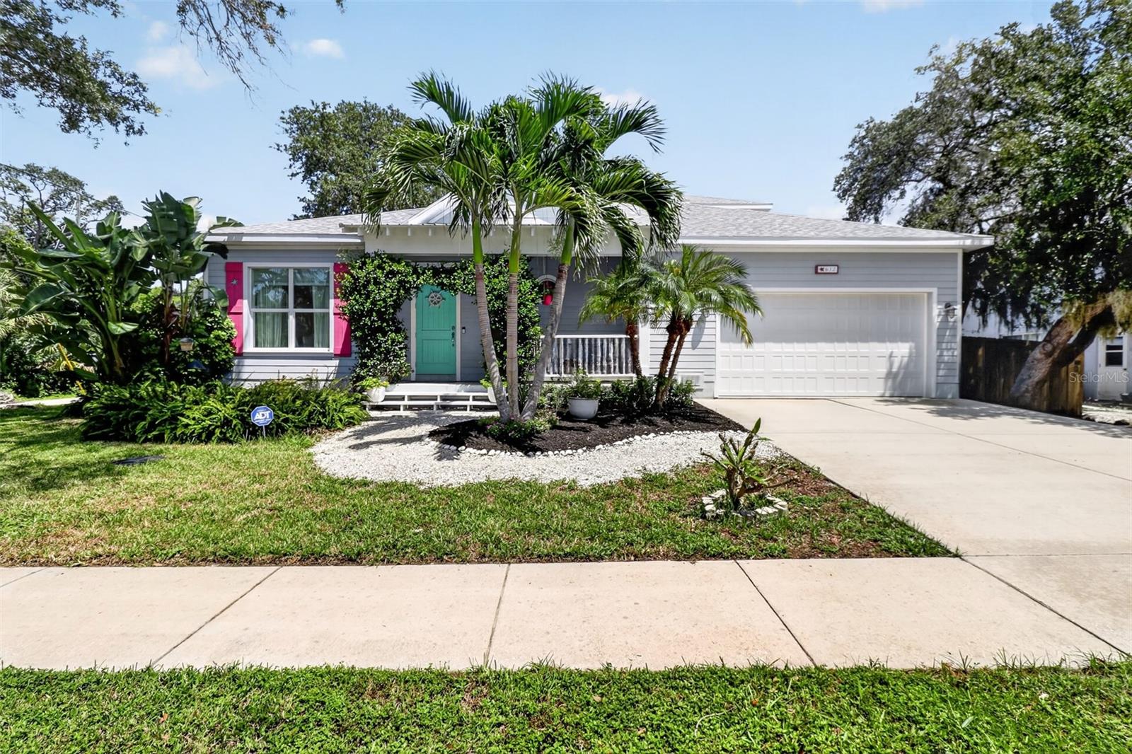 Gray single-story house with a teal front door, pink shutters, tropical landscaping, and an attached garage.