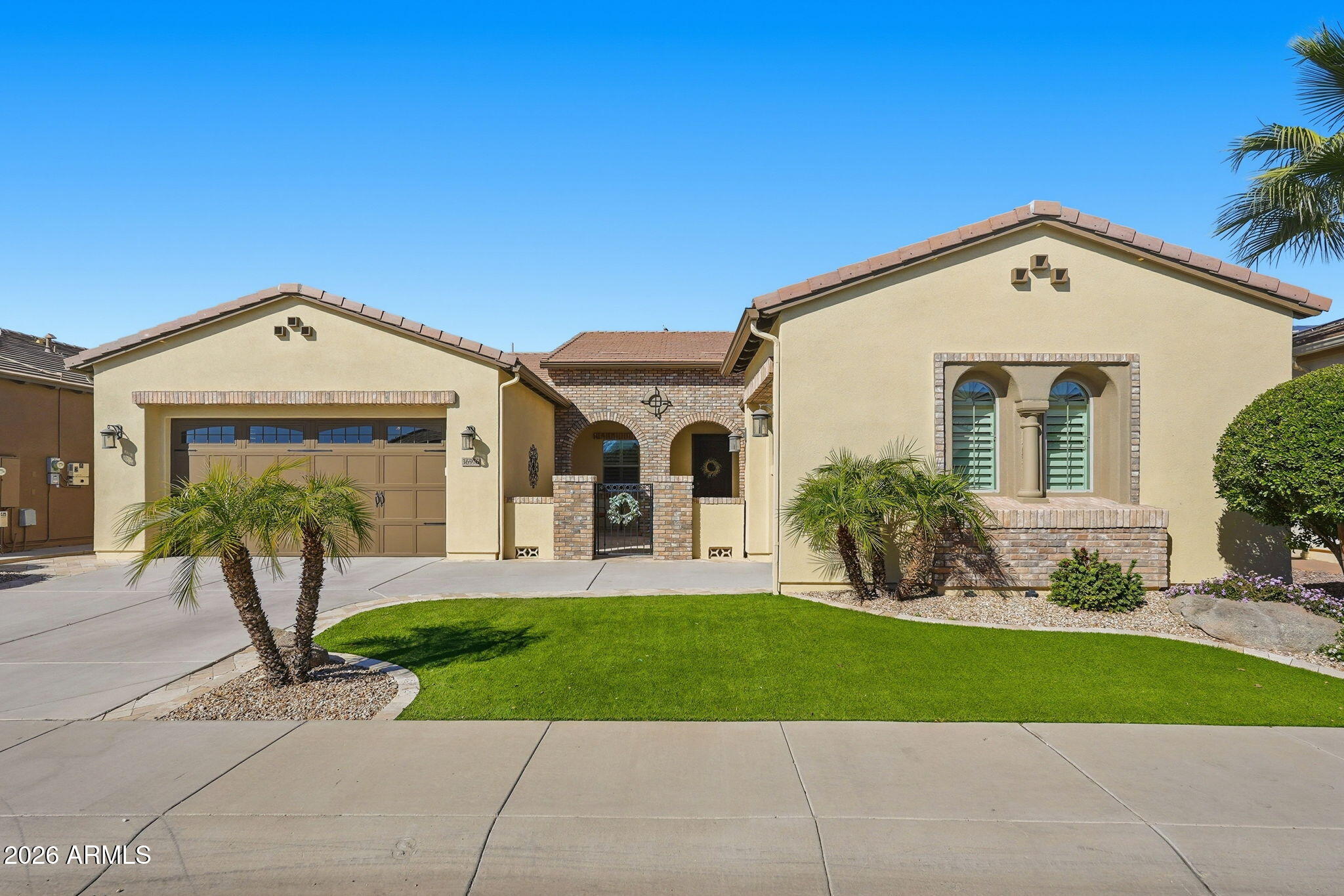 Beige stucco home with brick entry arches, a two-car garage, palm trees, and a curved lawn.