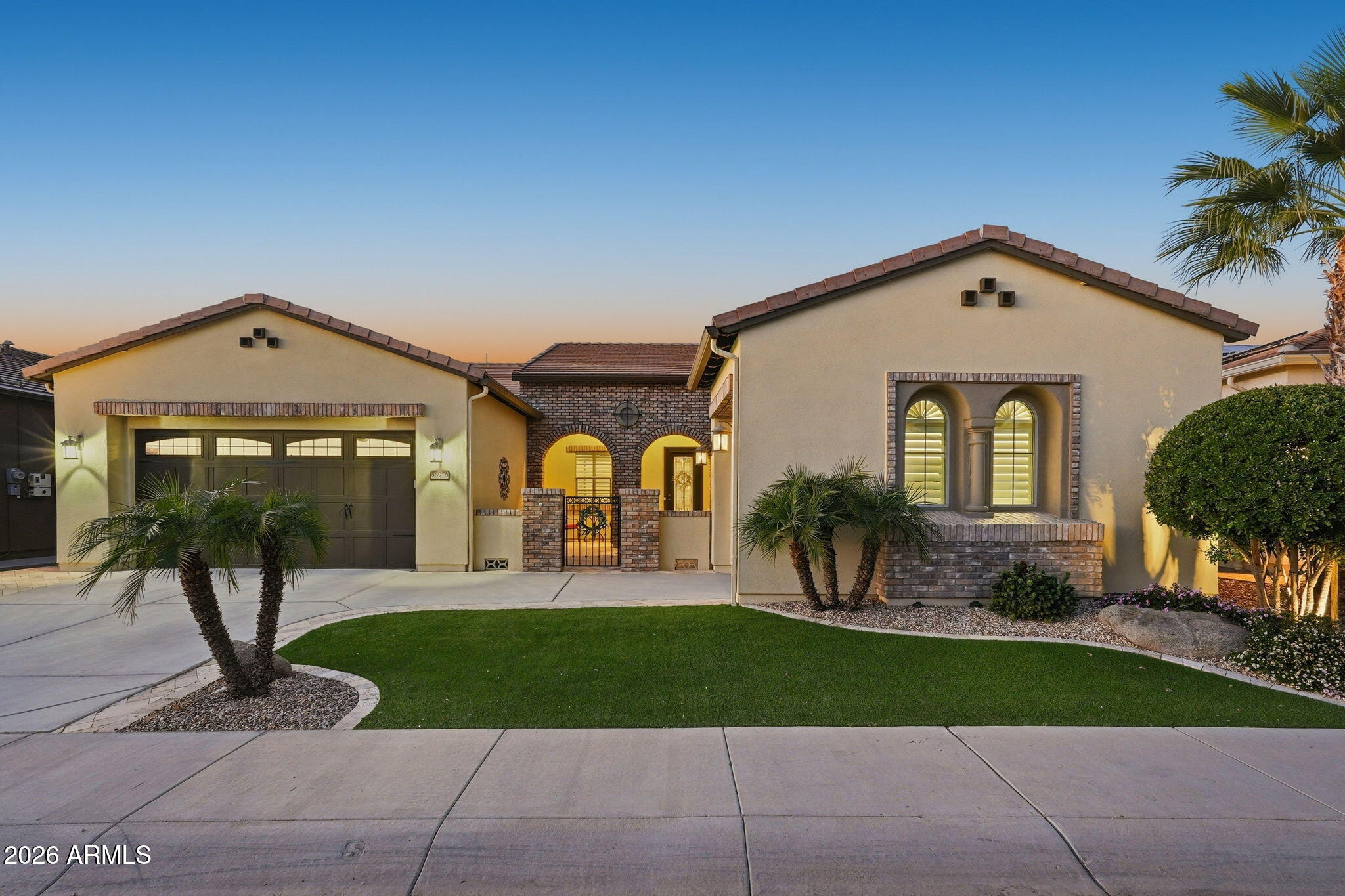 Stucco home with a two-car garage, brick entry, palm trees, and a curved green lawn at sunset.