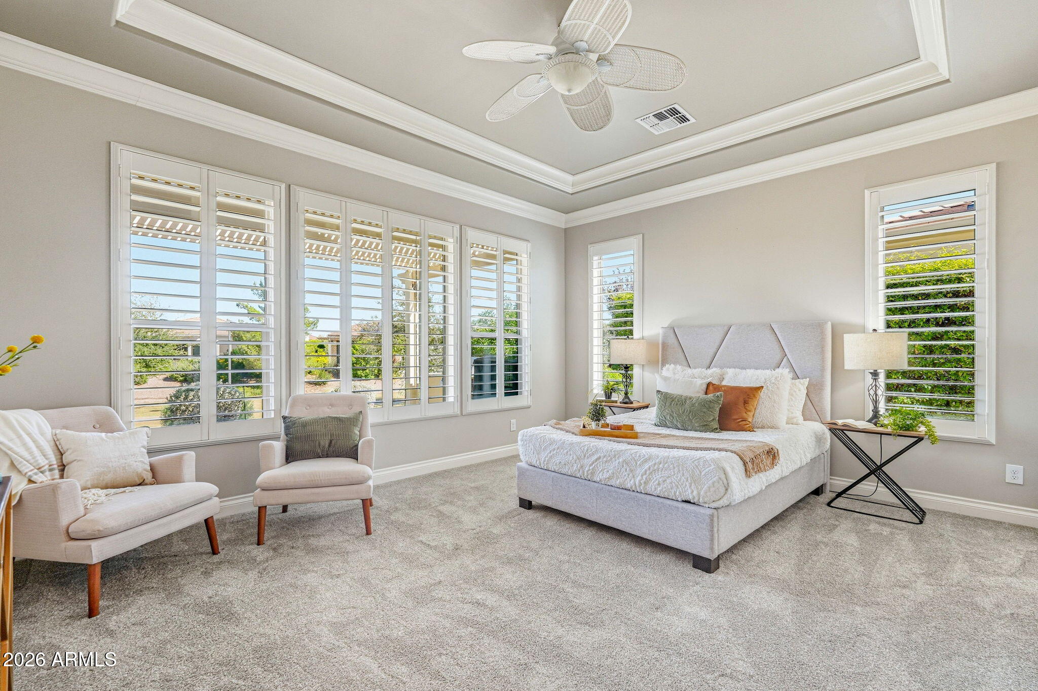 Staged bedroom with a king bed, two armchairs, and wide plantation-shuttered windows.
