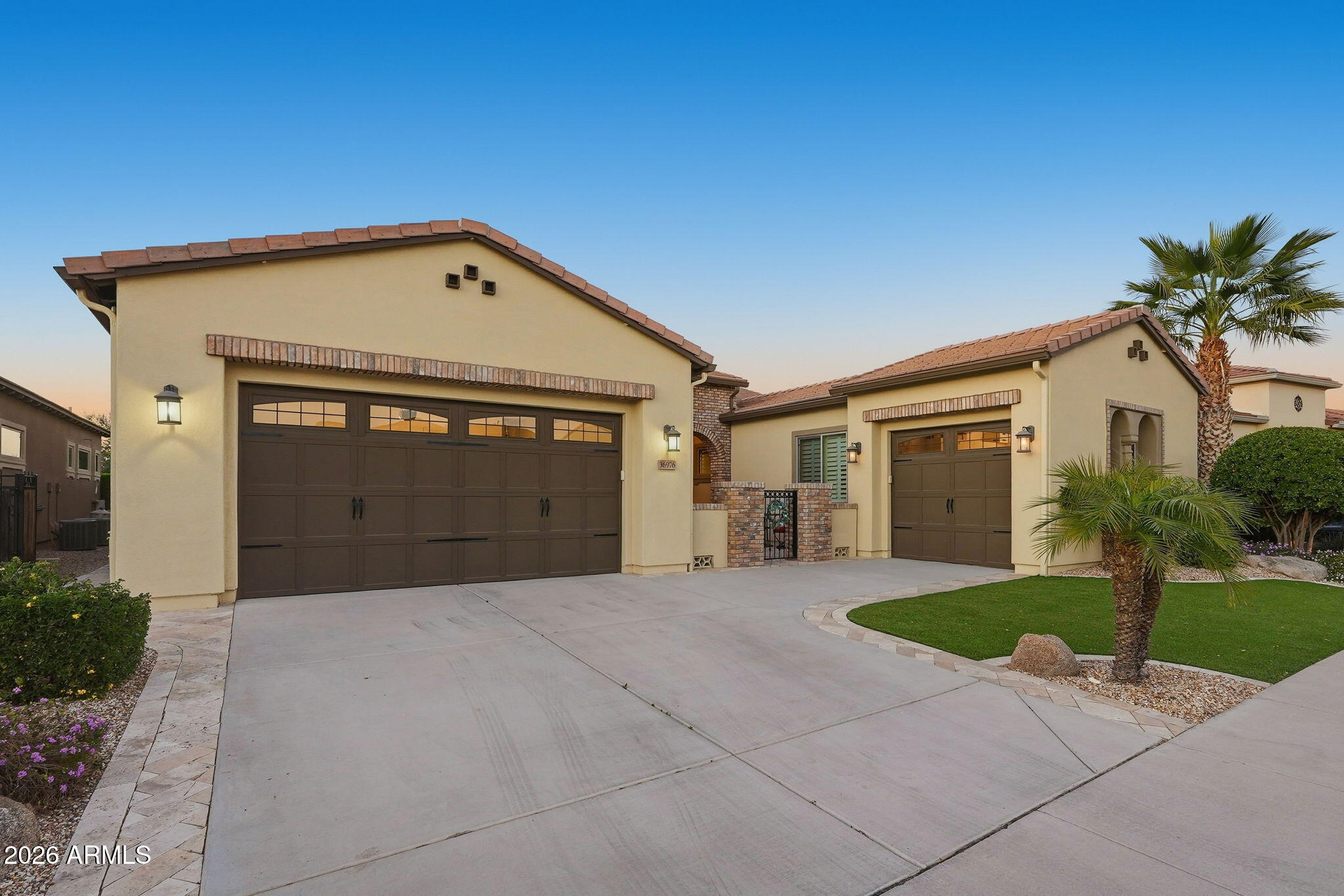 Beige stucco home with two dark garage doors, a wide driveway, and palm trees.