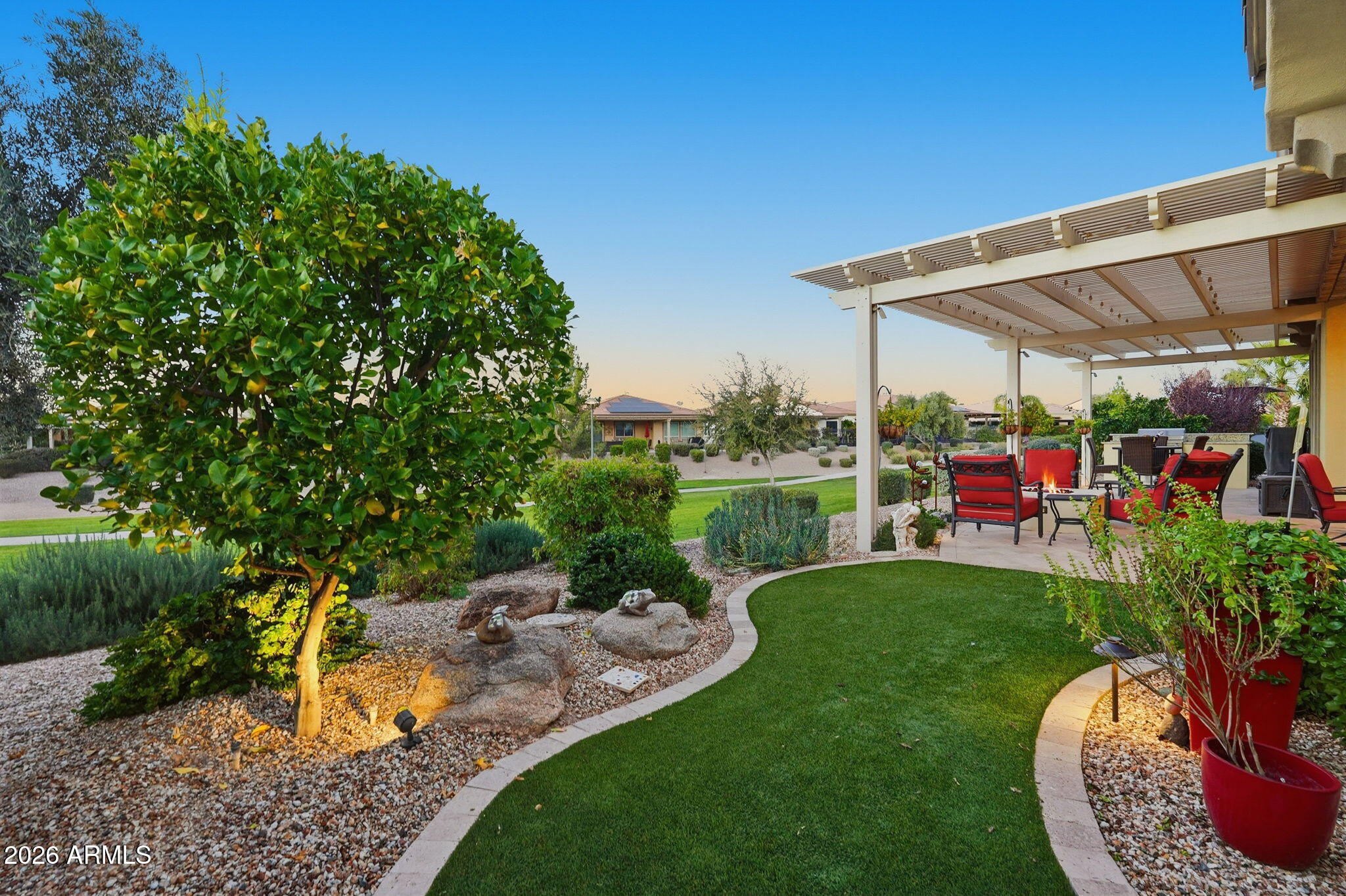 Landscaped backyard with a curved turf lawn and covered patio furnished with red chairs.