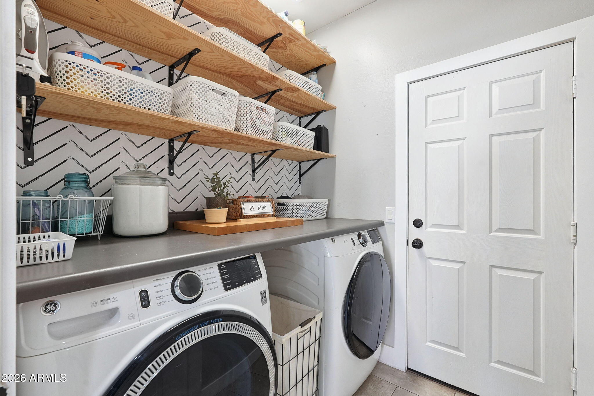 Laundry room with front-loading washer and dryer, wooden shelves, white baskets, and chevron wallpaper.